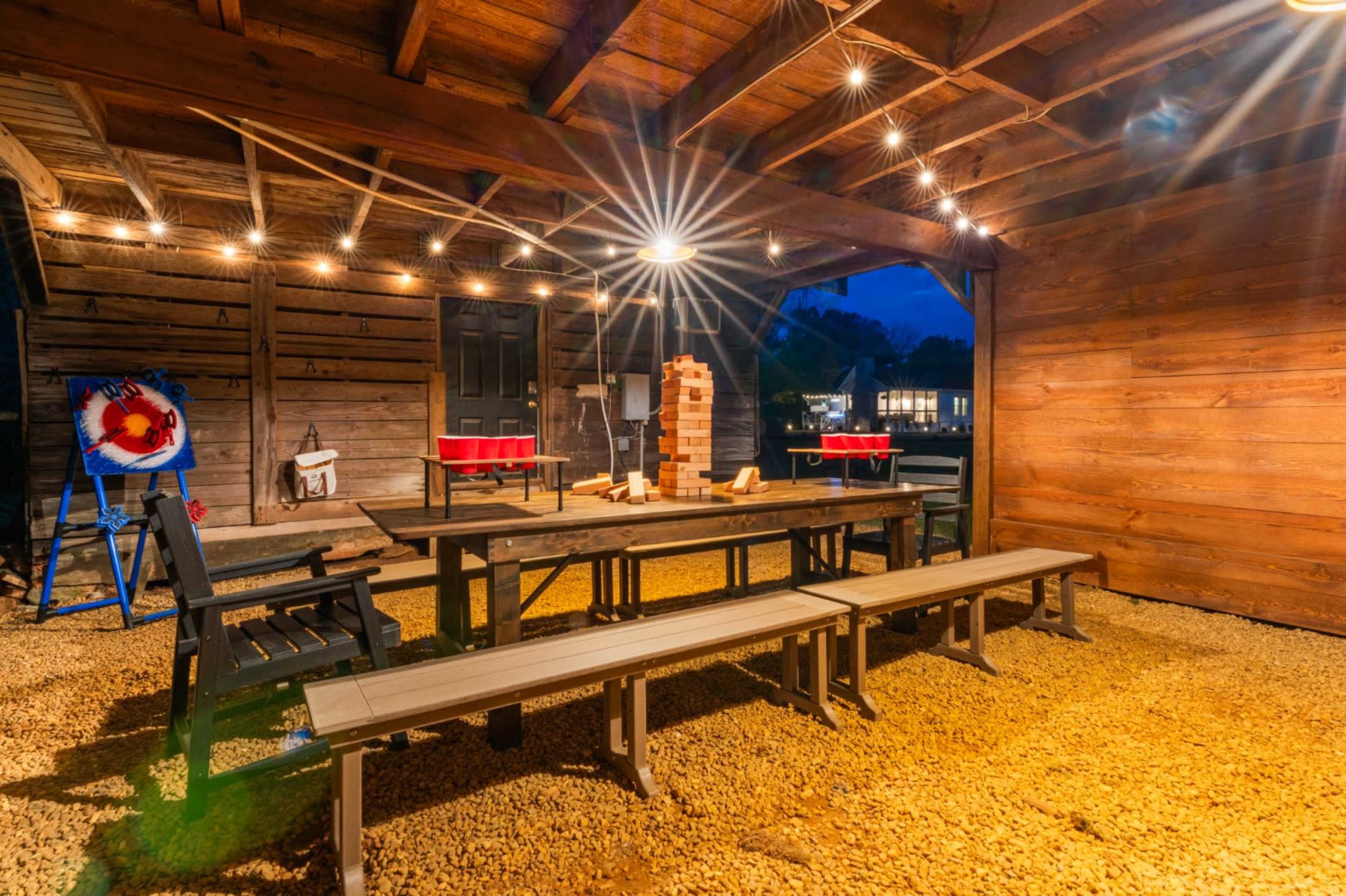 The image shows an outdoor seating area with a large table and benches beneath a wooden structure, illuminated by string lights.