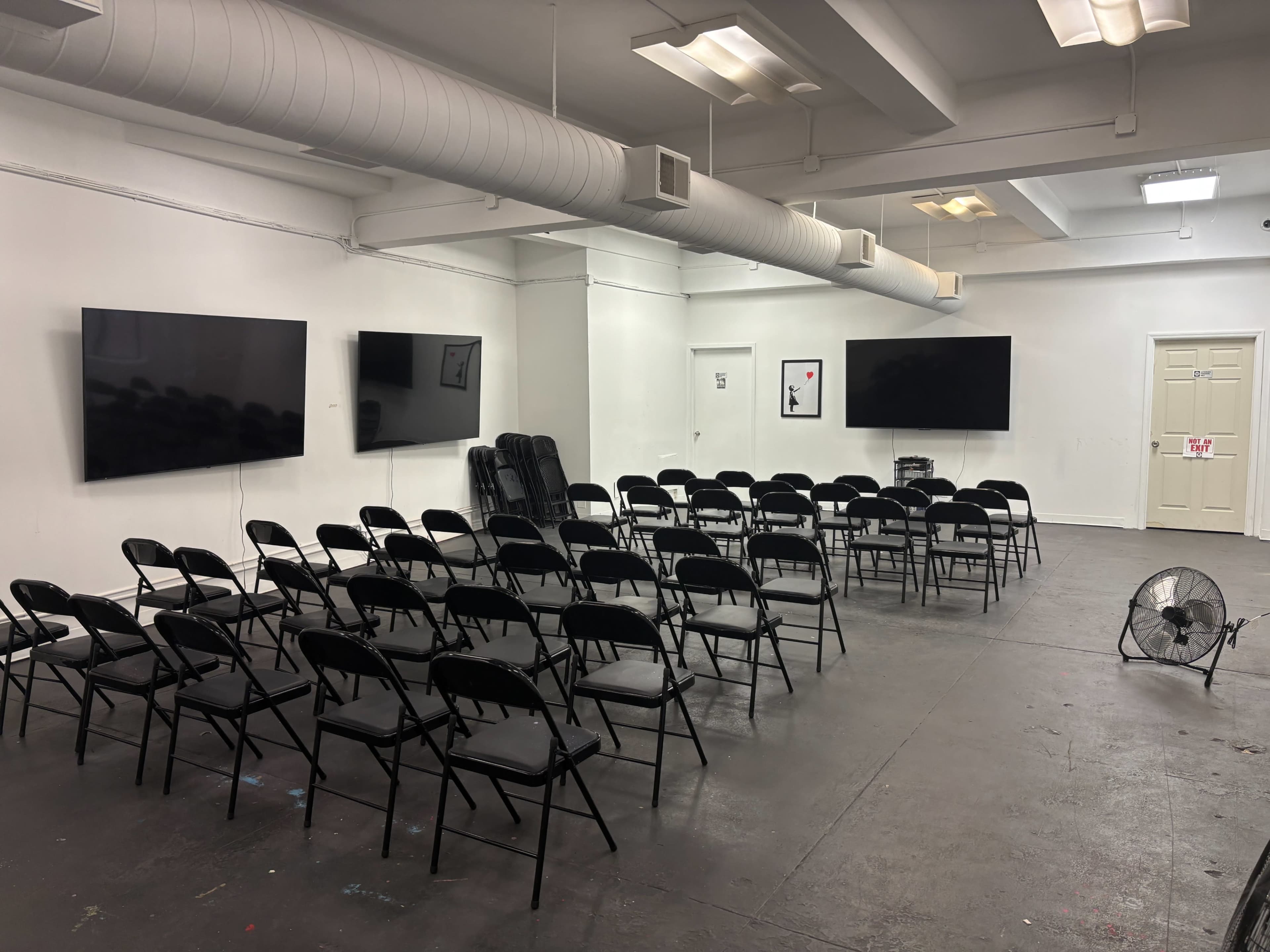 The image shows a spacious room set up for a presentation, featuring several rows of black chairs facing large flat-screen televisions mounted on the walls.