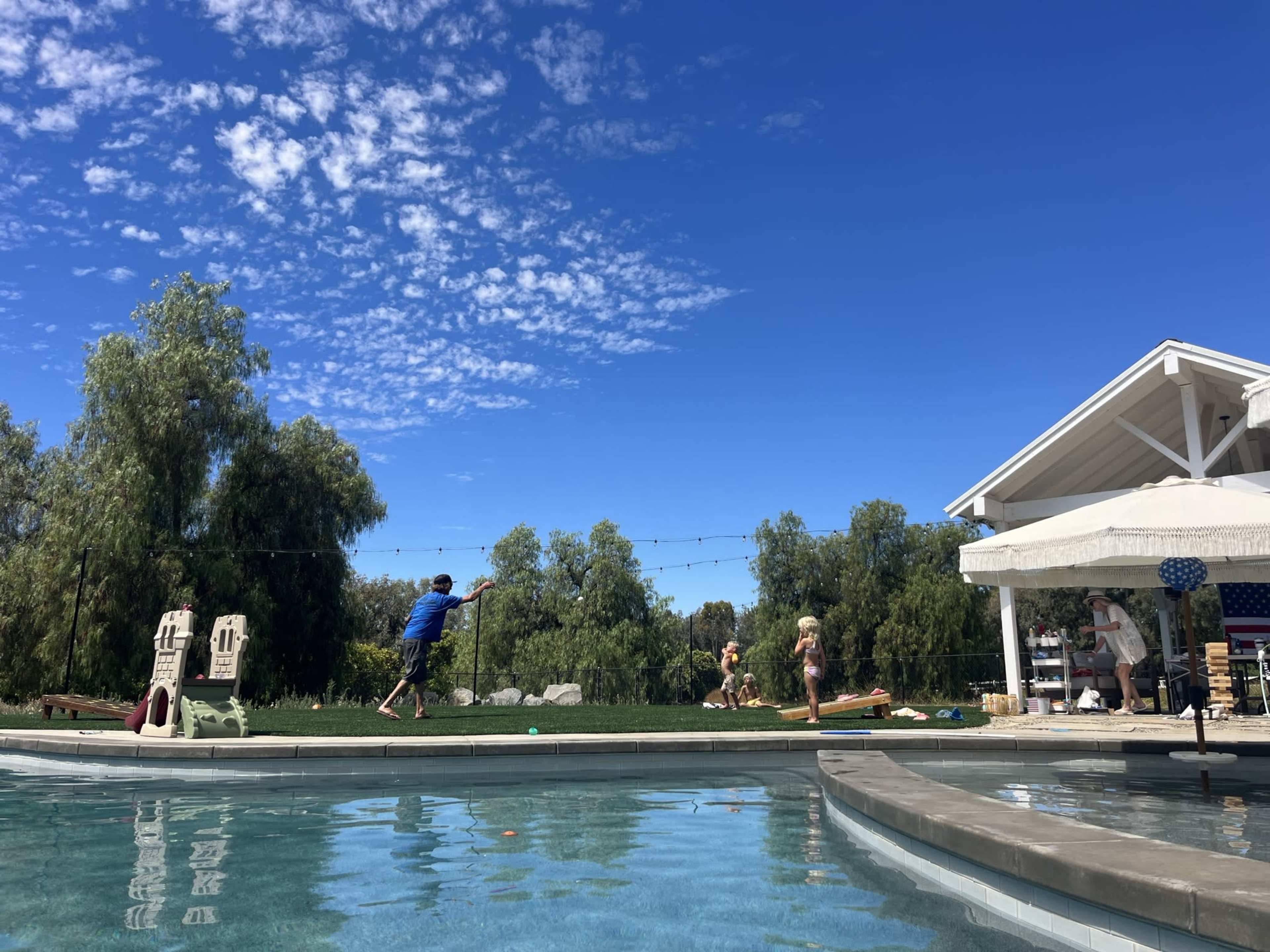 A boy in a blue shirt plays near a pool while two children engage in an activity on the grass beside a white gazebo under a clear blue sky.