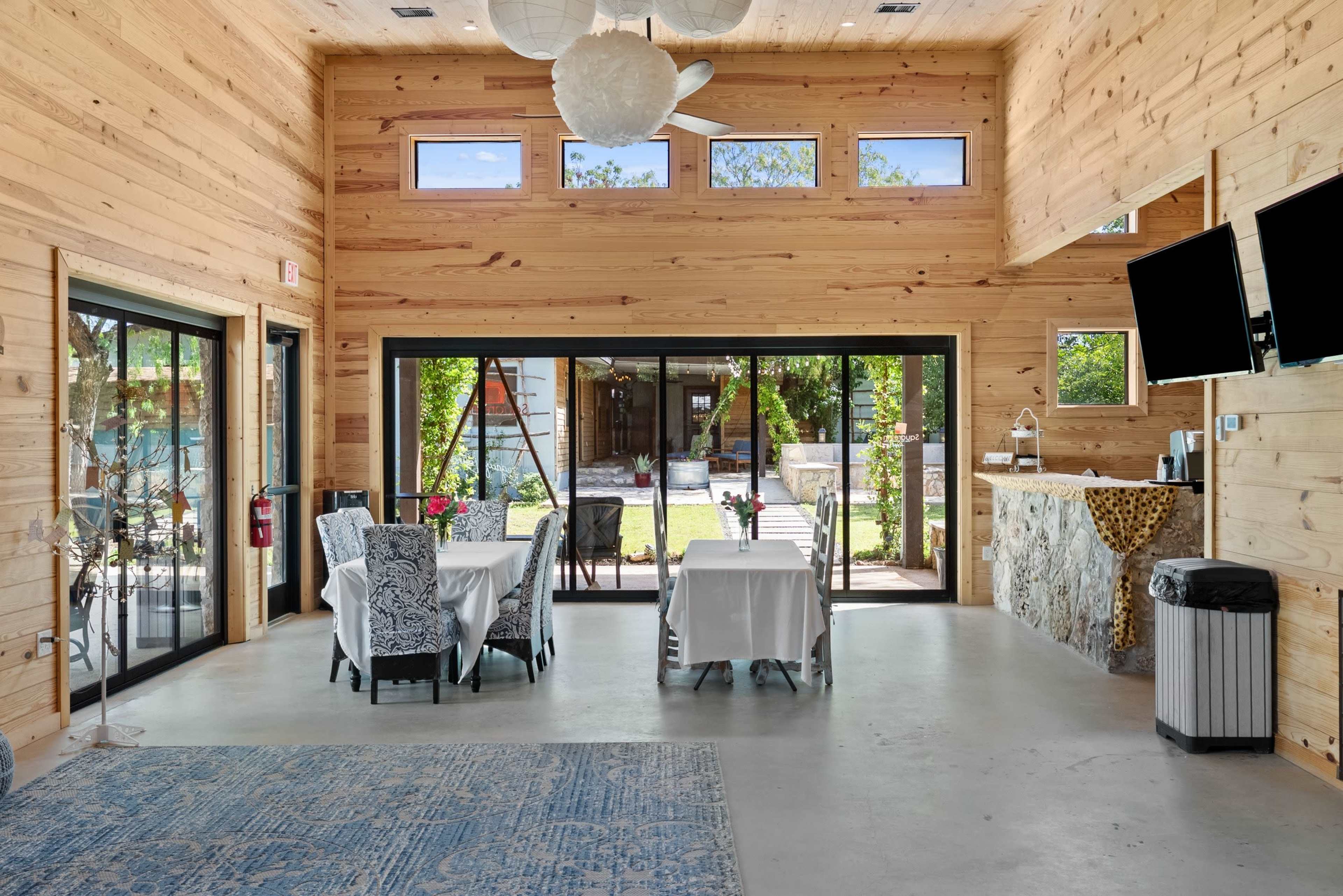 The interior of a wooden dining area features two tables set for meals, with large glass doors opening to a garden space outside.