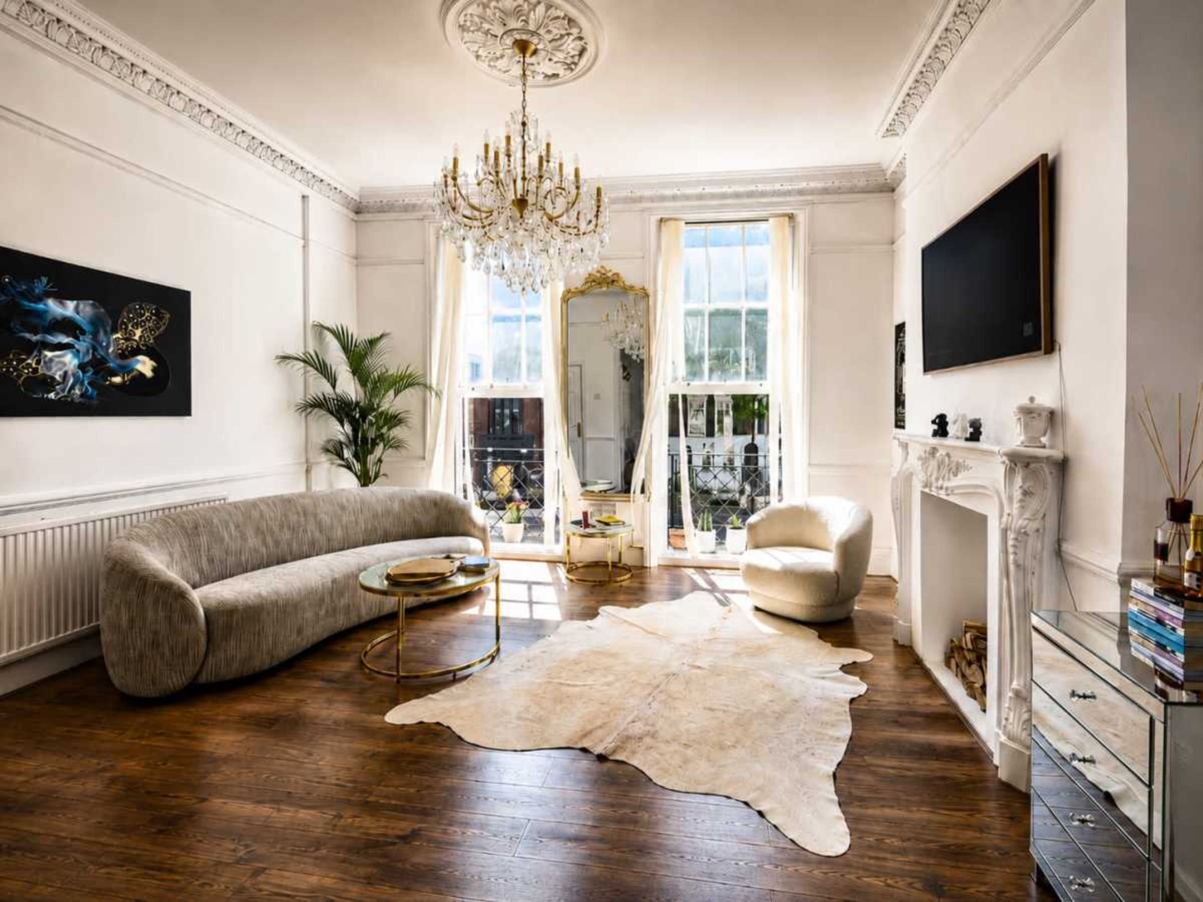 The living room features a curved sofa, a glass coffee table, a decorative chandelier, and large windows providing natural light, with a rug positioned on the wooden floor.