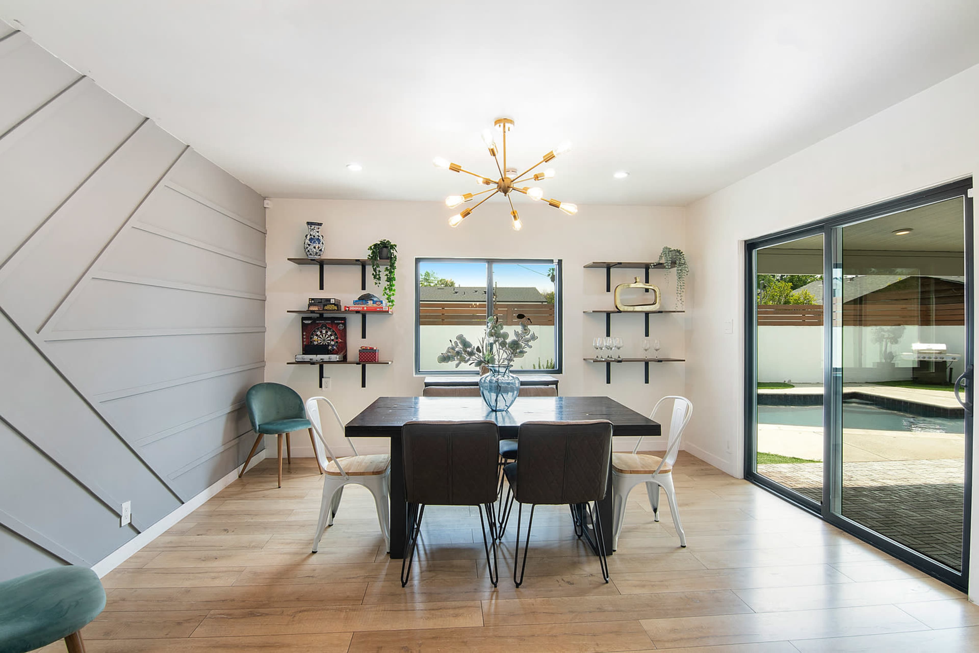 The image showcases a modern dining room featuring a wooden table surrounded by six chairs, with decorative shelves and a large window overlooking a pool area.