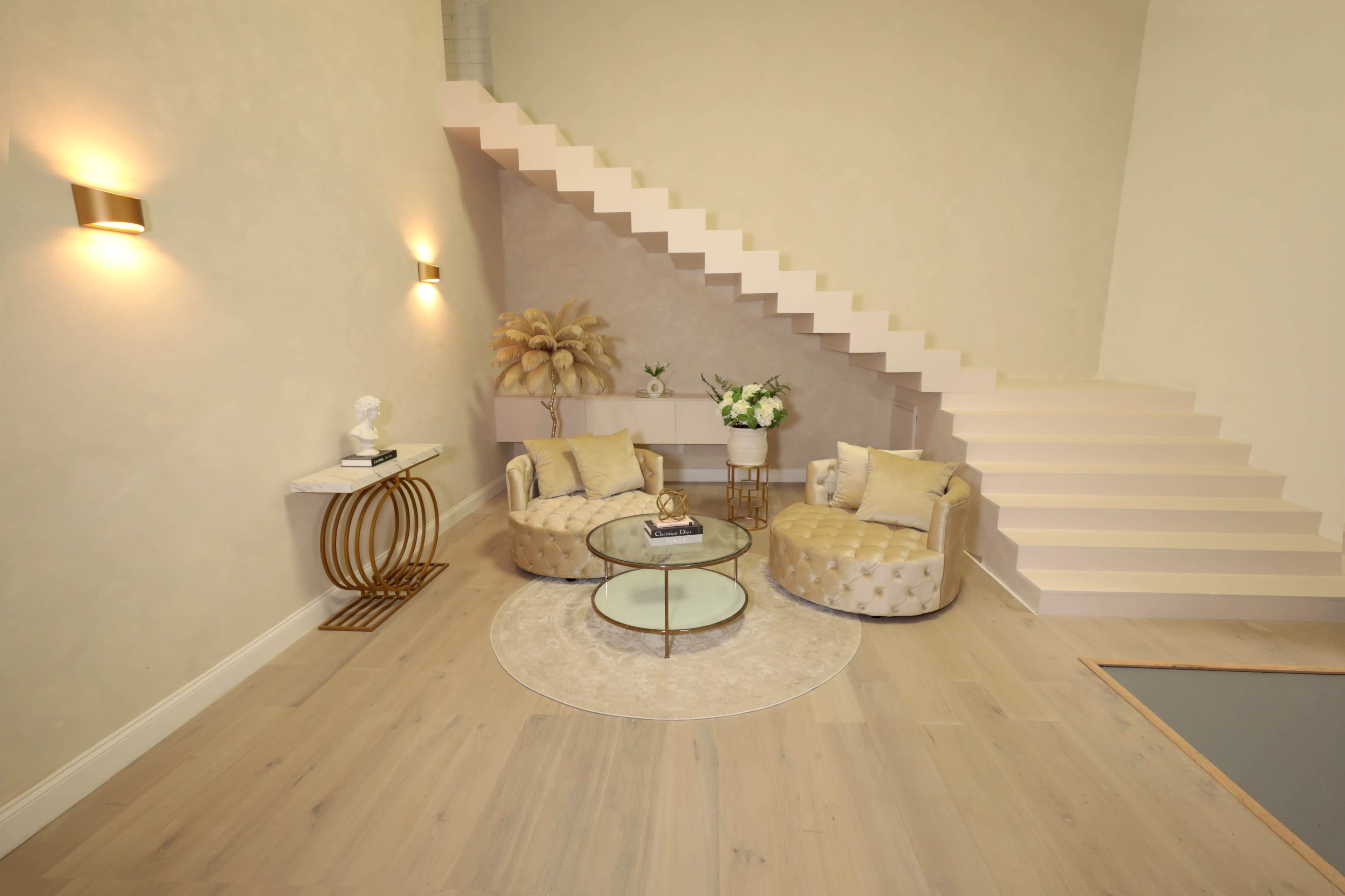 A modern interior with two tufted chairs, a round glass coffee table, and a staircase leading to an upper level.