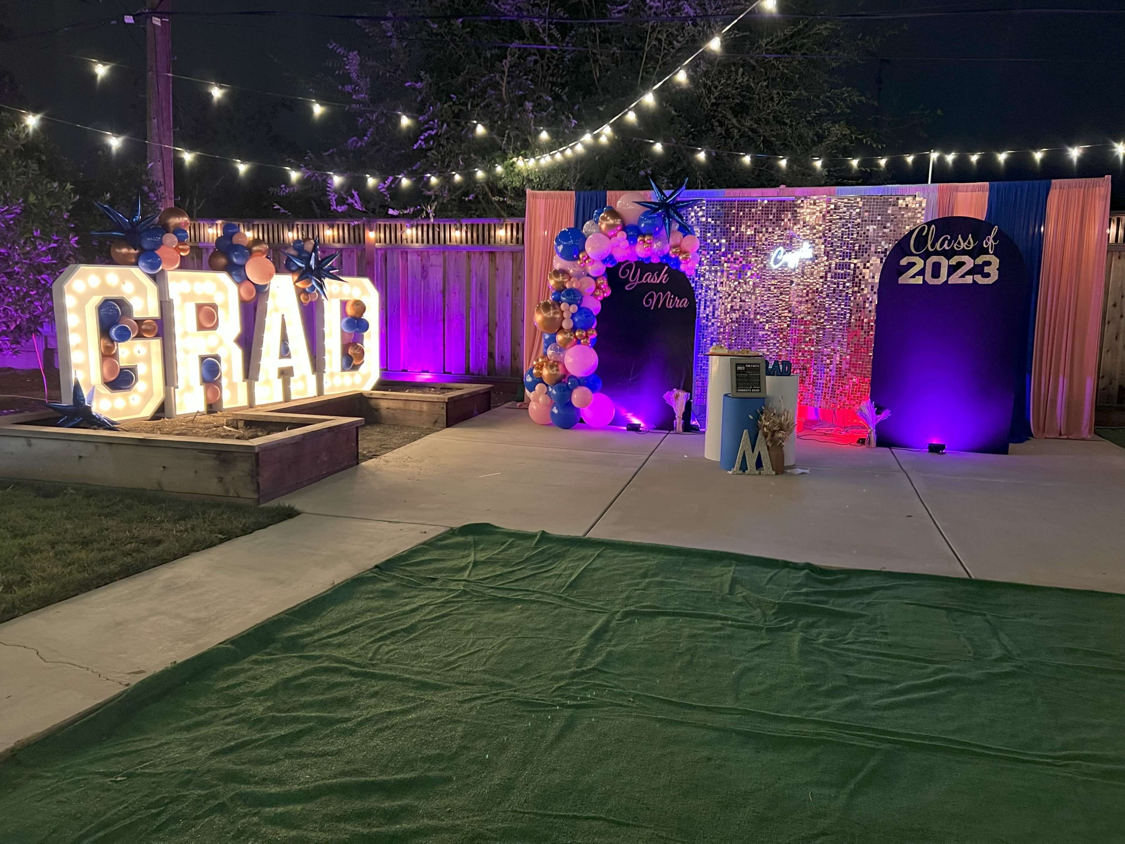 The scene features a decorated outdoor space for a graduation celebration, including illuminated letters spelling "GRAD," a backdrop with "Class of 2023," and colorful balloons.