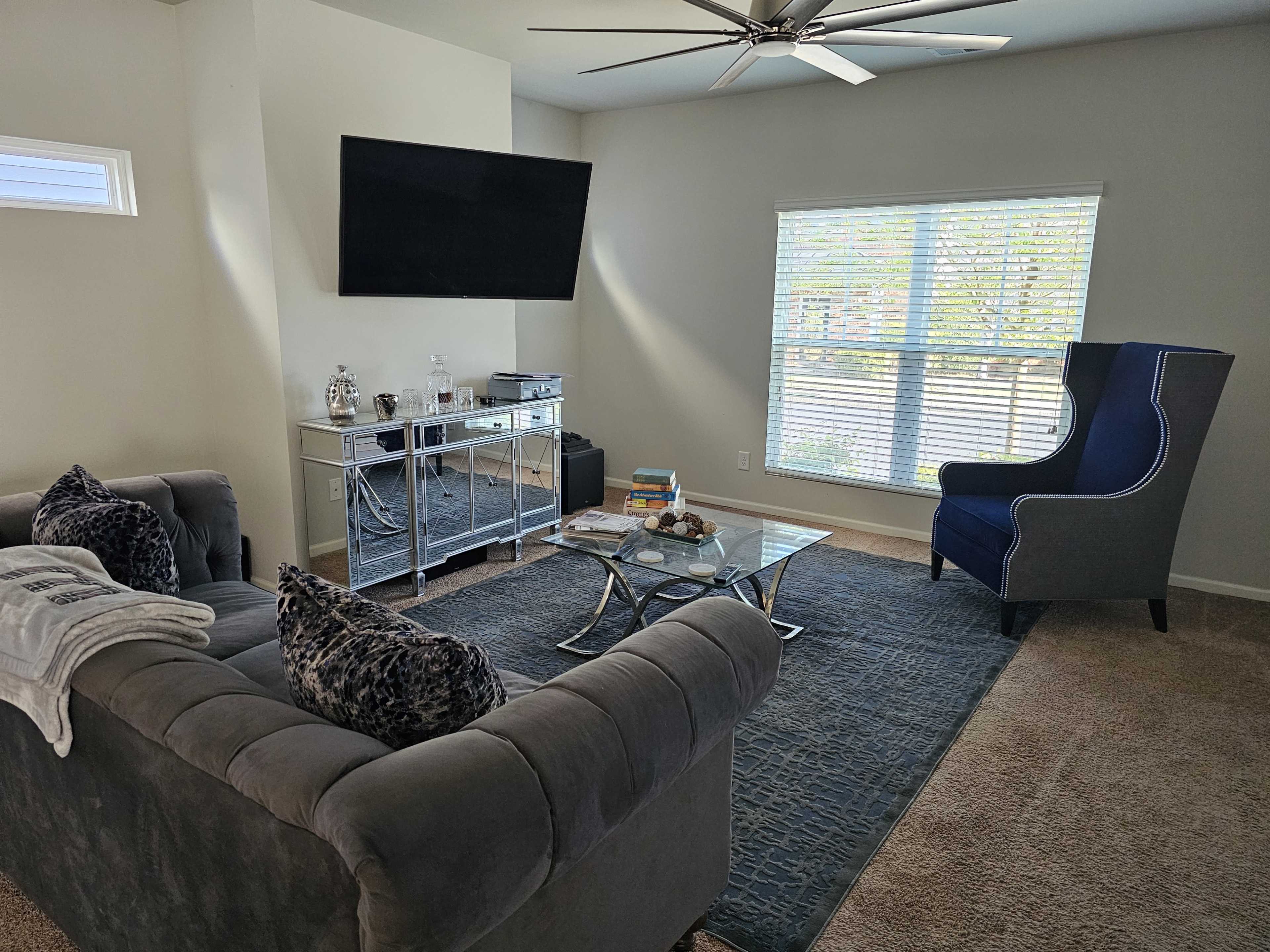 A living room with a gray sectional sofa, a glass coffee table, a large TV mounted on the wall, and a blue accent chair next to a window with blinds.