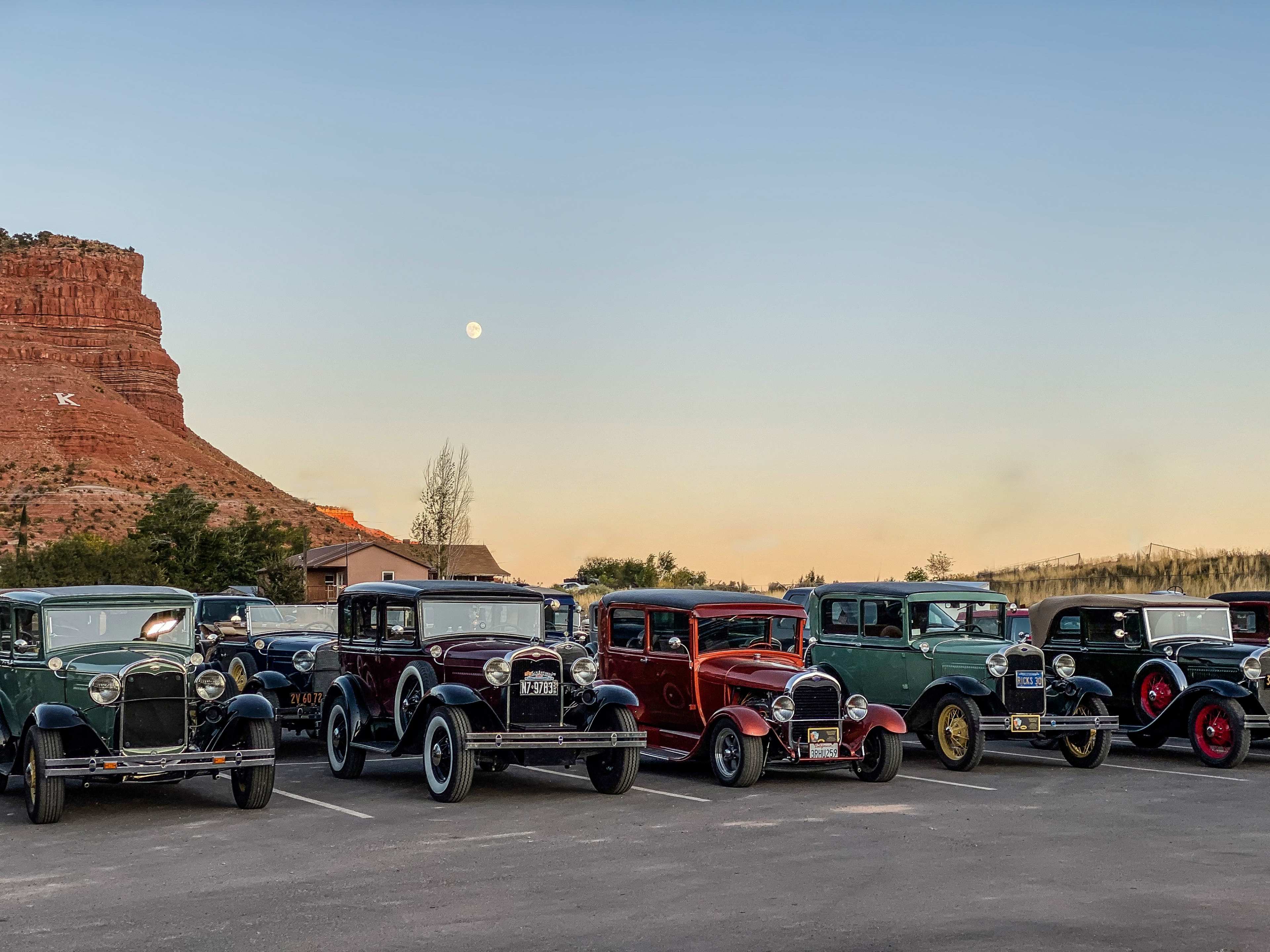 A line of vintage cars from the 1920s is parked in a lot with a rocky cliff and a full moon in the background.