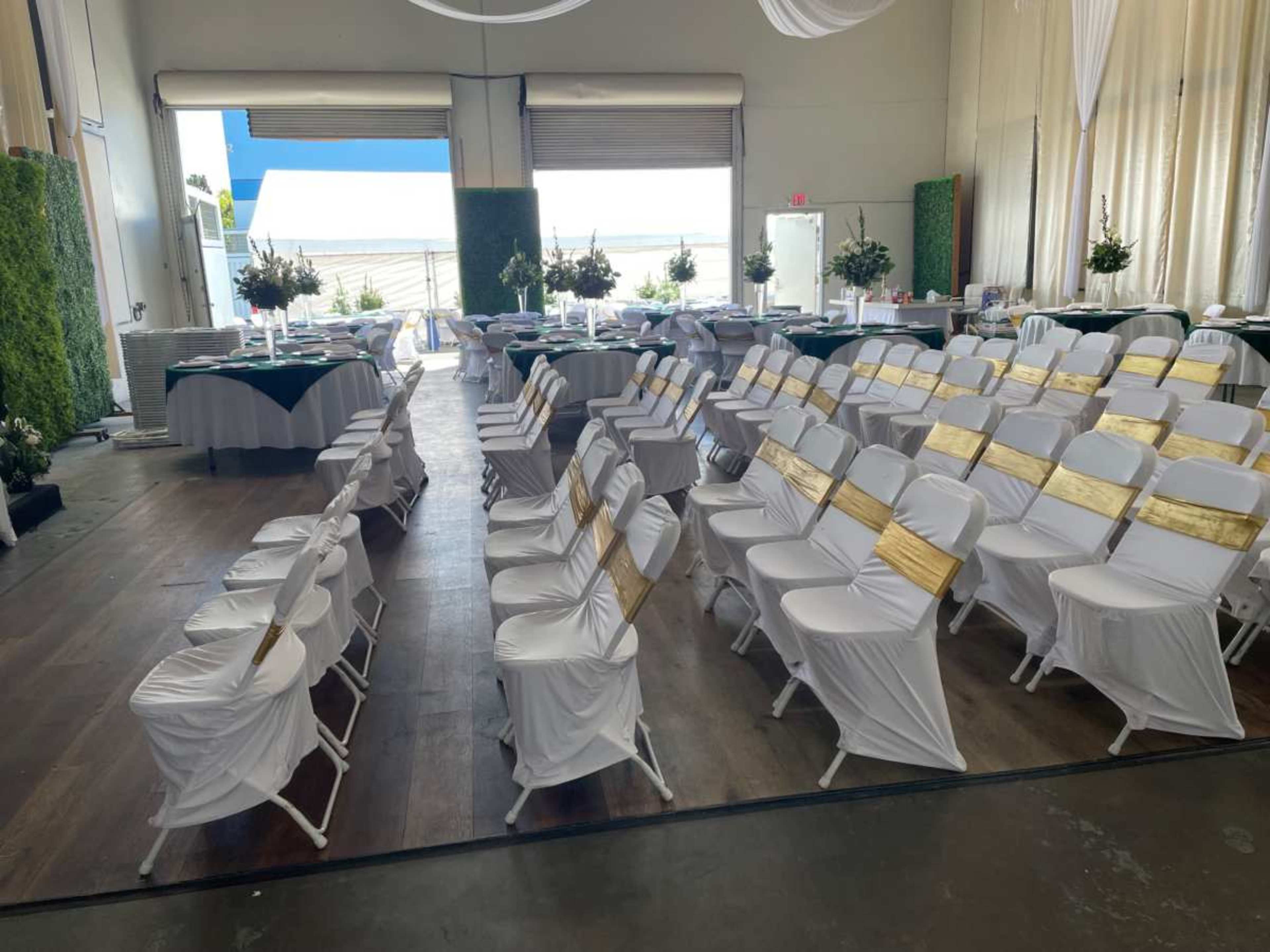 A spacious indoor venue is arranged for an event, featuring rows of white chairs with gold sashes and tables set with decorative centerpieces.
