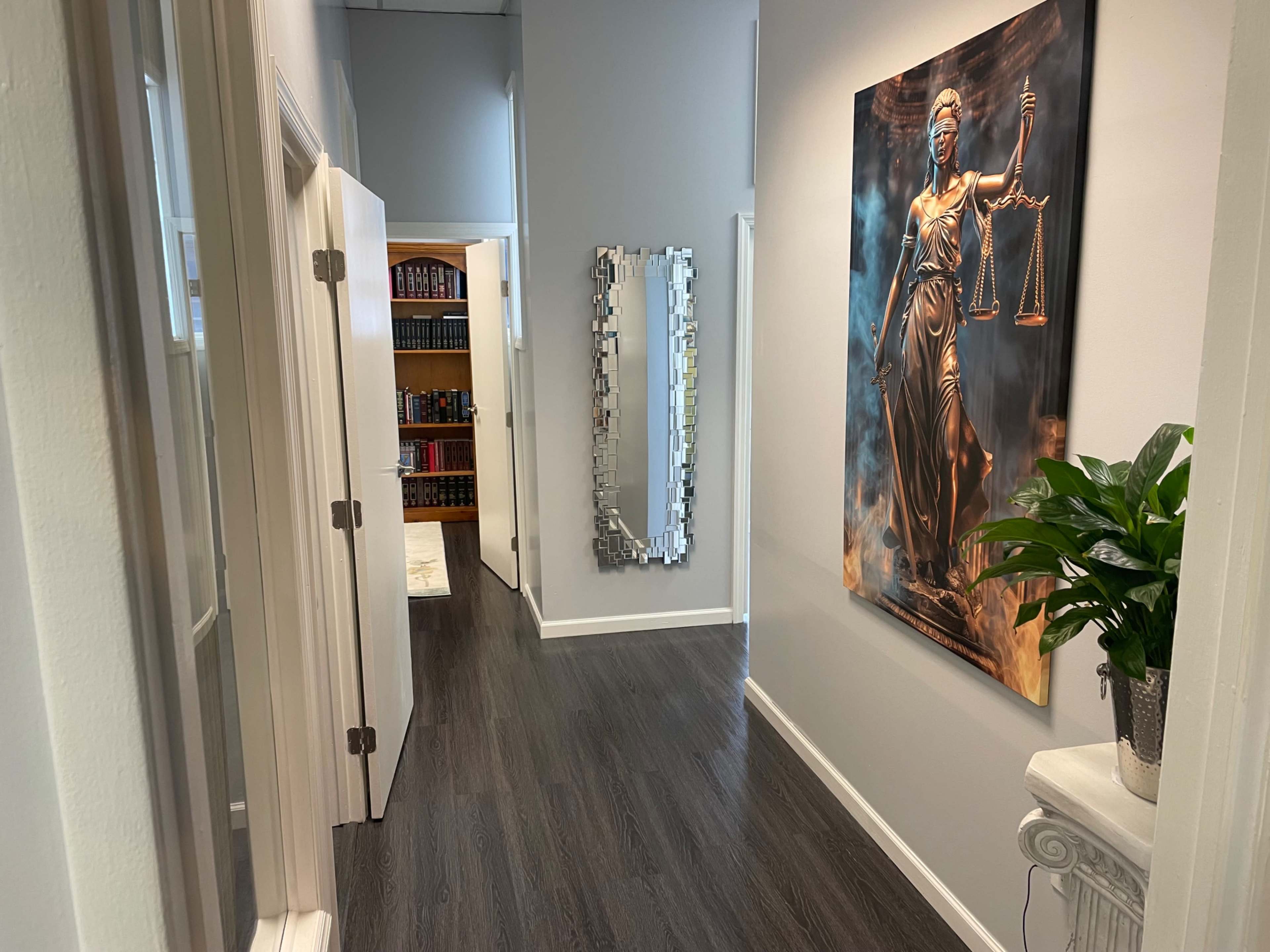 A well-lit hallway features a large statue of Lady Justice on the wall, white doors on either side, a mirrored sculpture, and a bookshelf in the background.