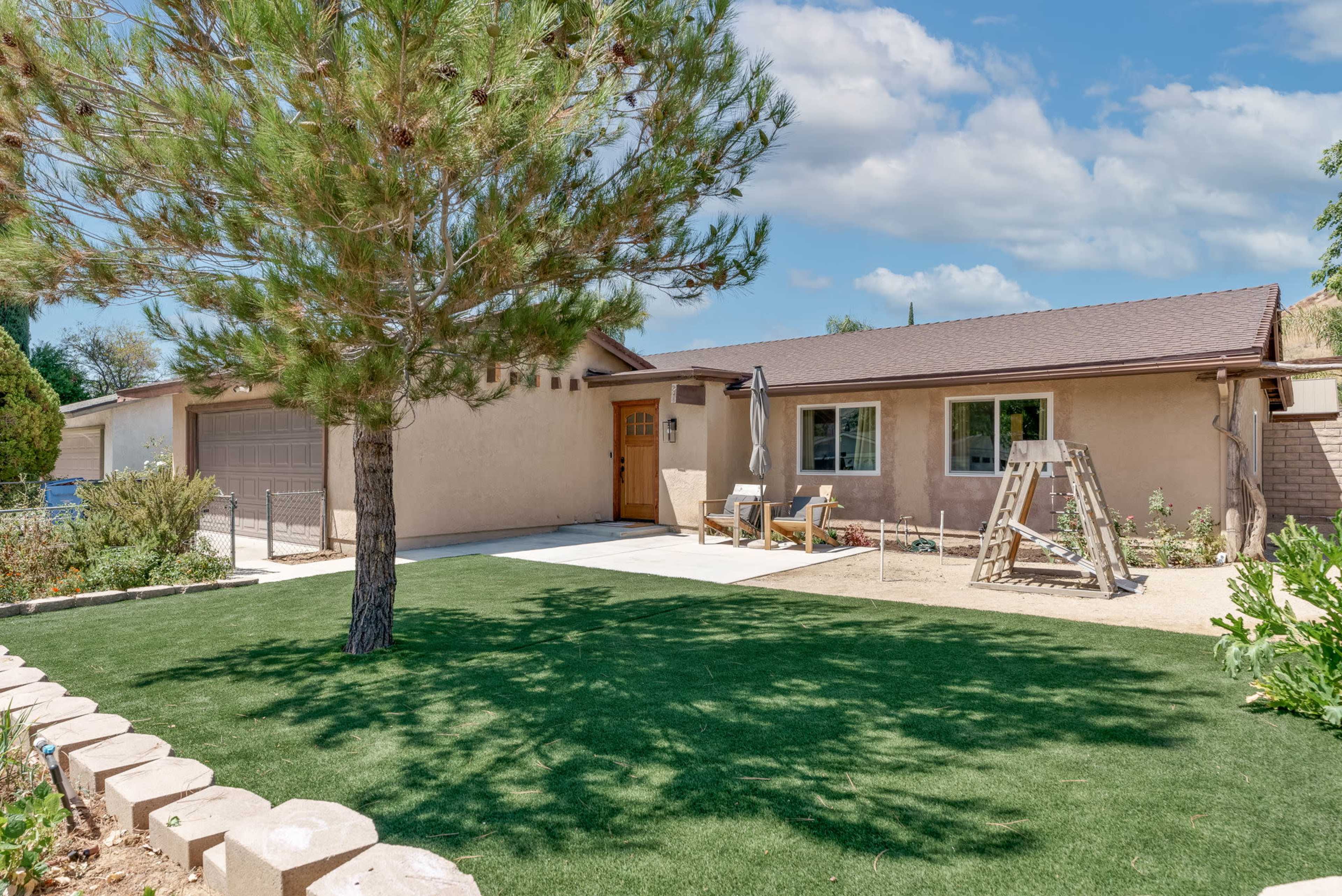 The image shows a single-story house with a brown roof, a driveway leading to a garage, and a front yard featuring a large tree and manicured grass.