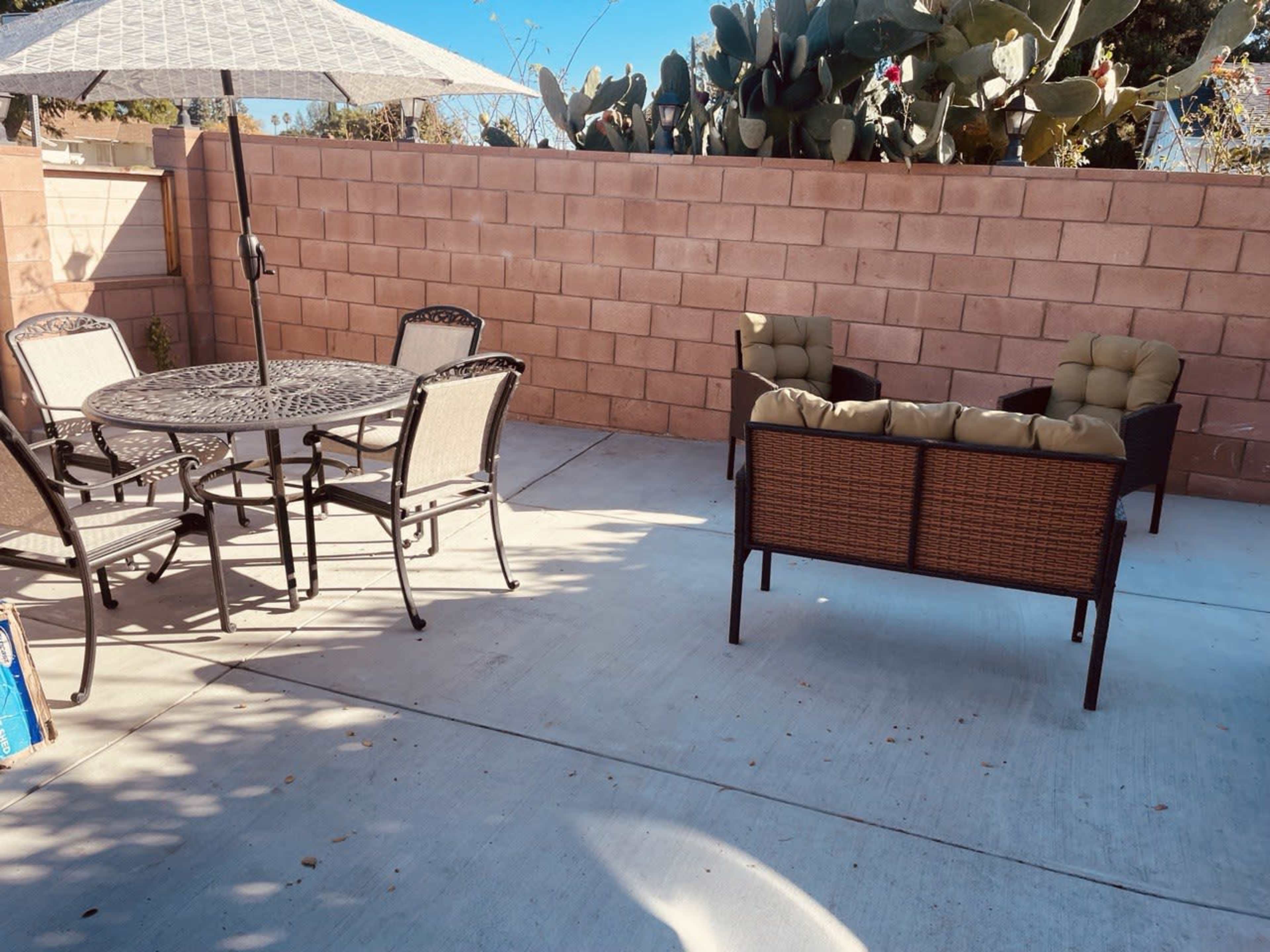 A patio area featuring a round table with six chairs, a lounge seating arrangement, and a large umbrella, surrounded by a stone wall and cacti.