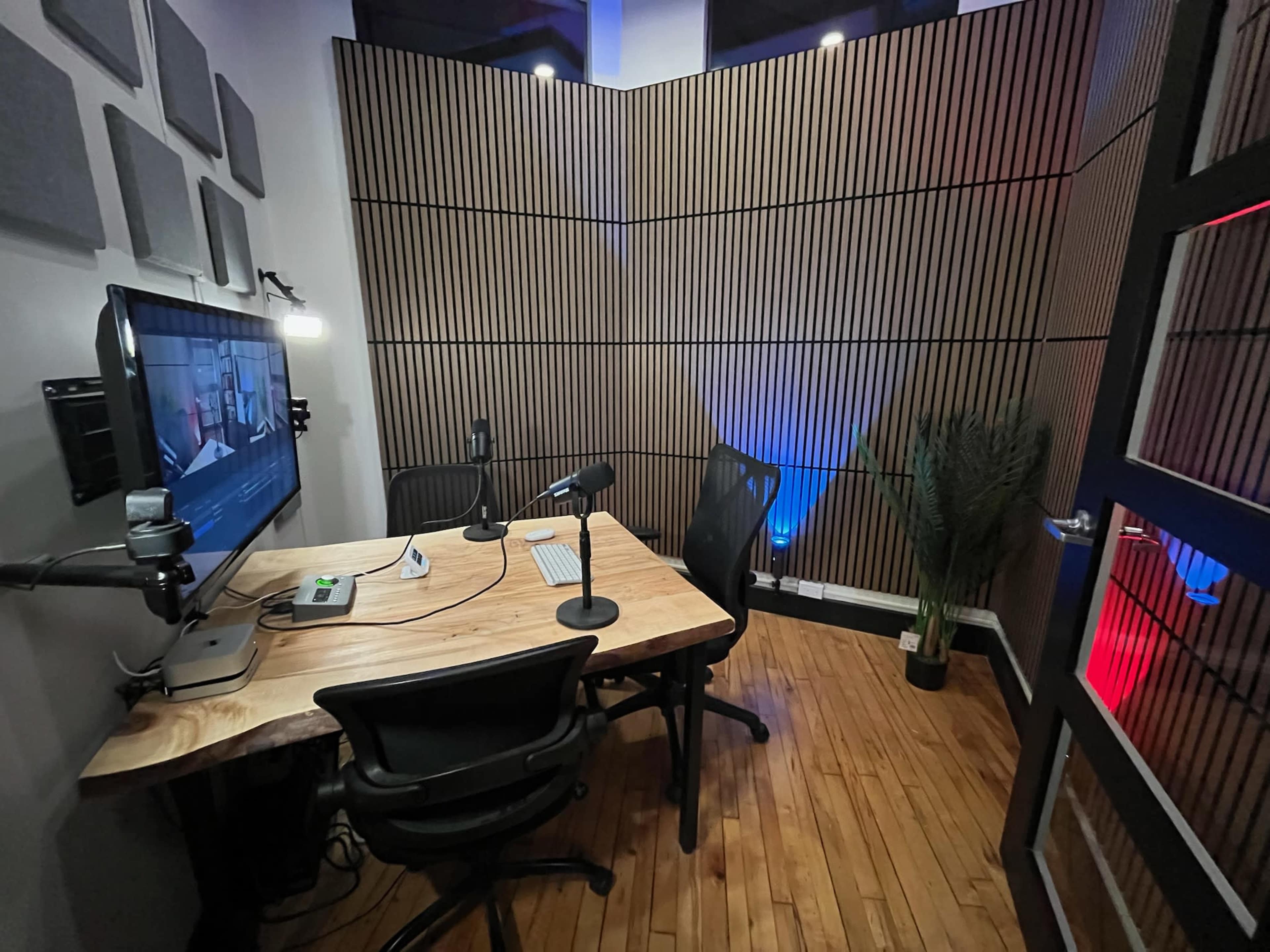 The image shows a small recording studio with wooden walls, a large monitor, two microphones on stands, and a minimalist wooden desk.