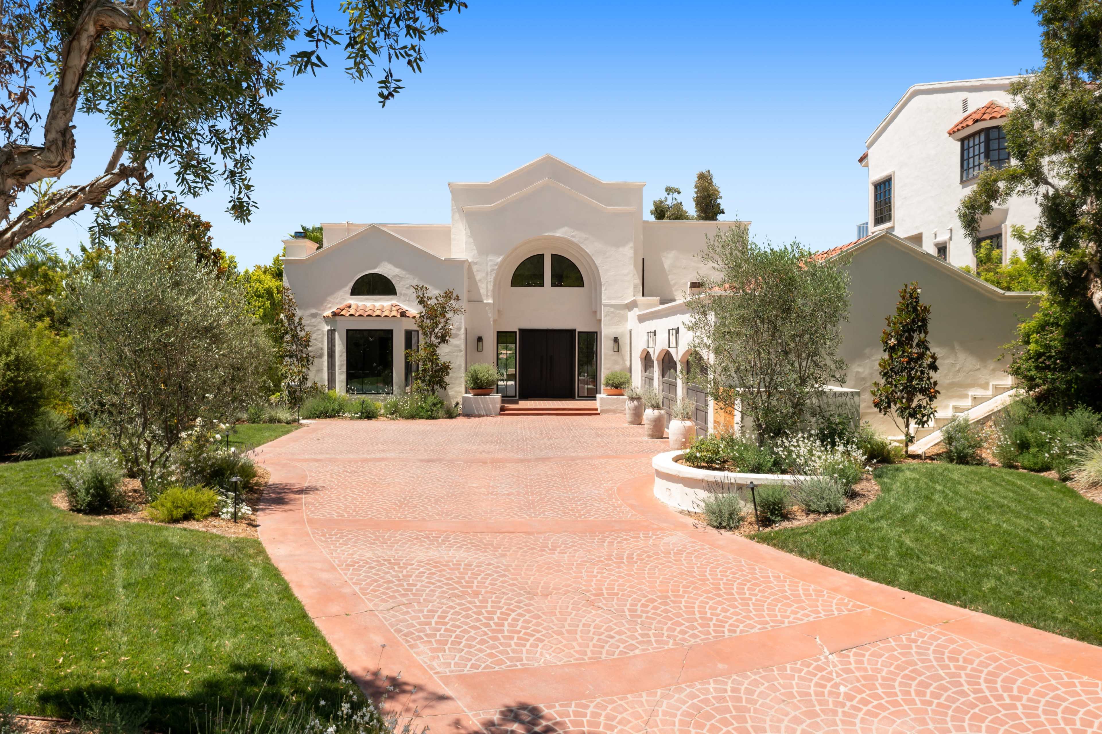 A large, white stucco house with a red brick driveway and landscaped greenery surrounding it.