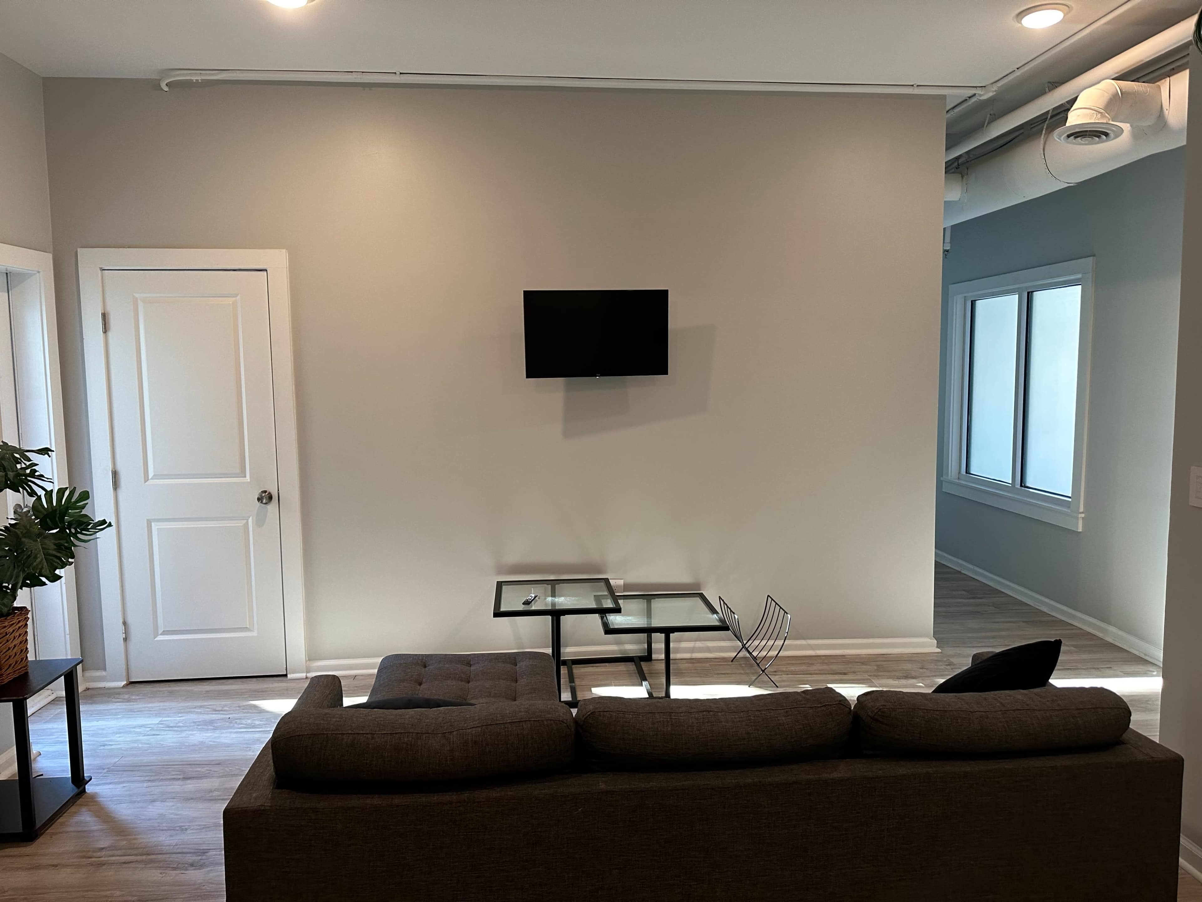 A gray sofa faces a glass coffee table, with a wall-mounted TV above it in a minimalistic living space featuring a door and windows.