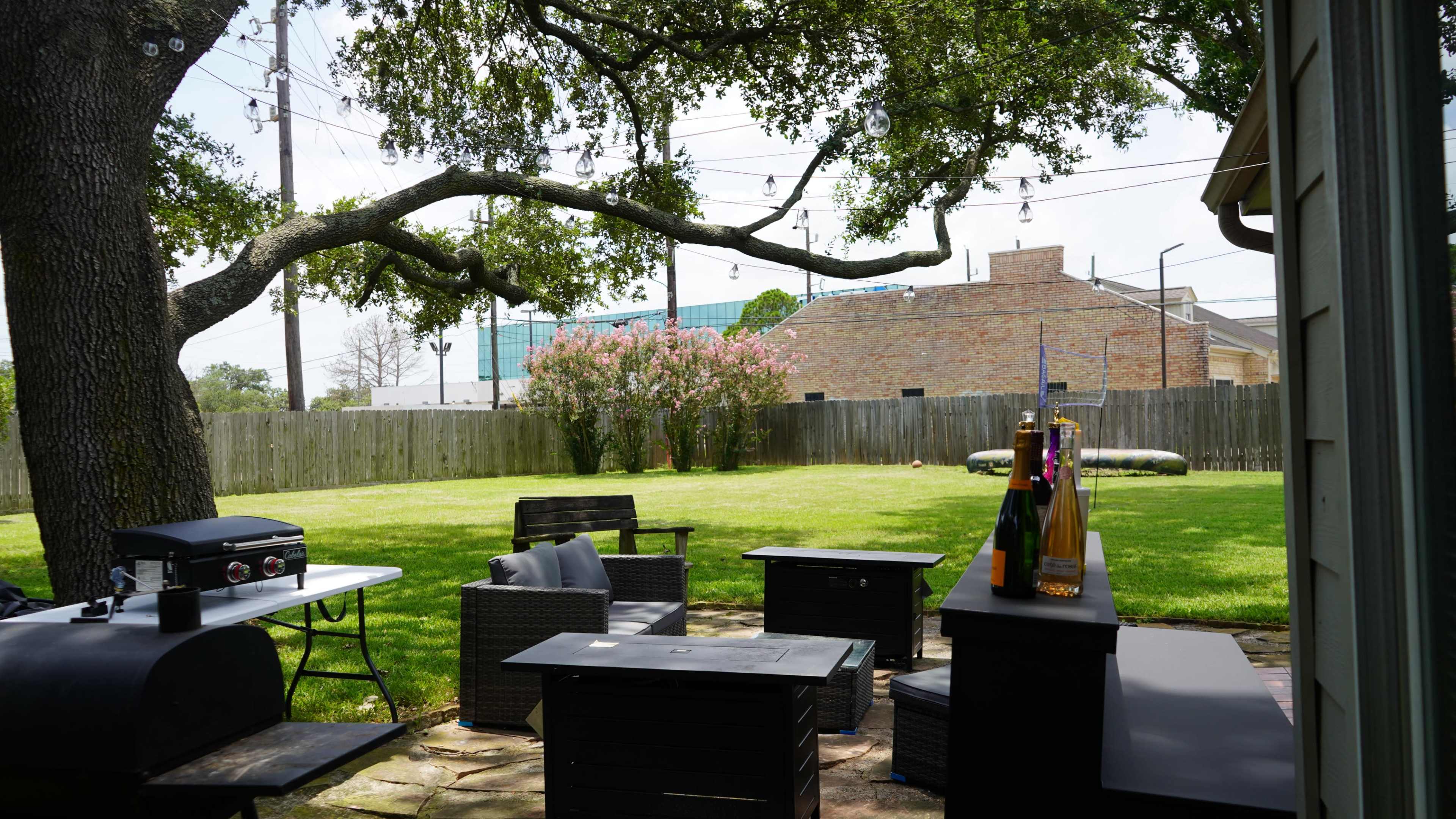 The image depicts a backyard with patio furniture, a grill, and a well-maintained lawn, framed by a large tree with hanging lights.