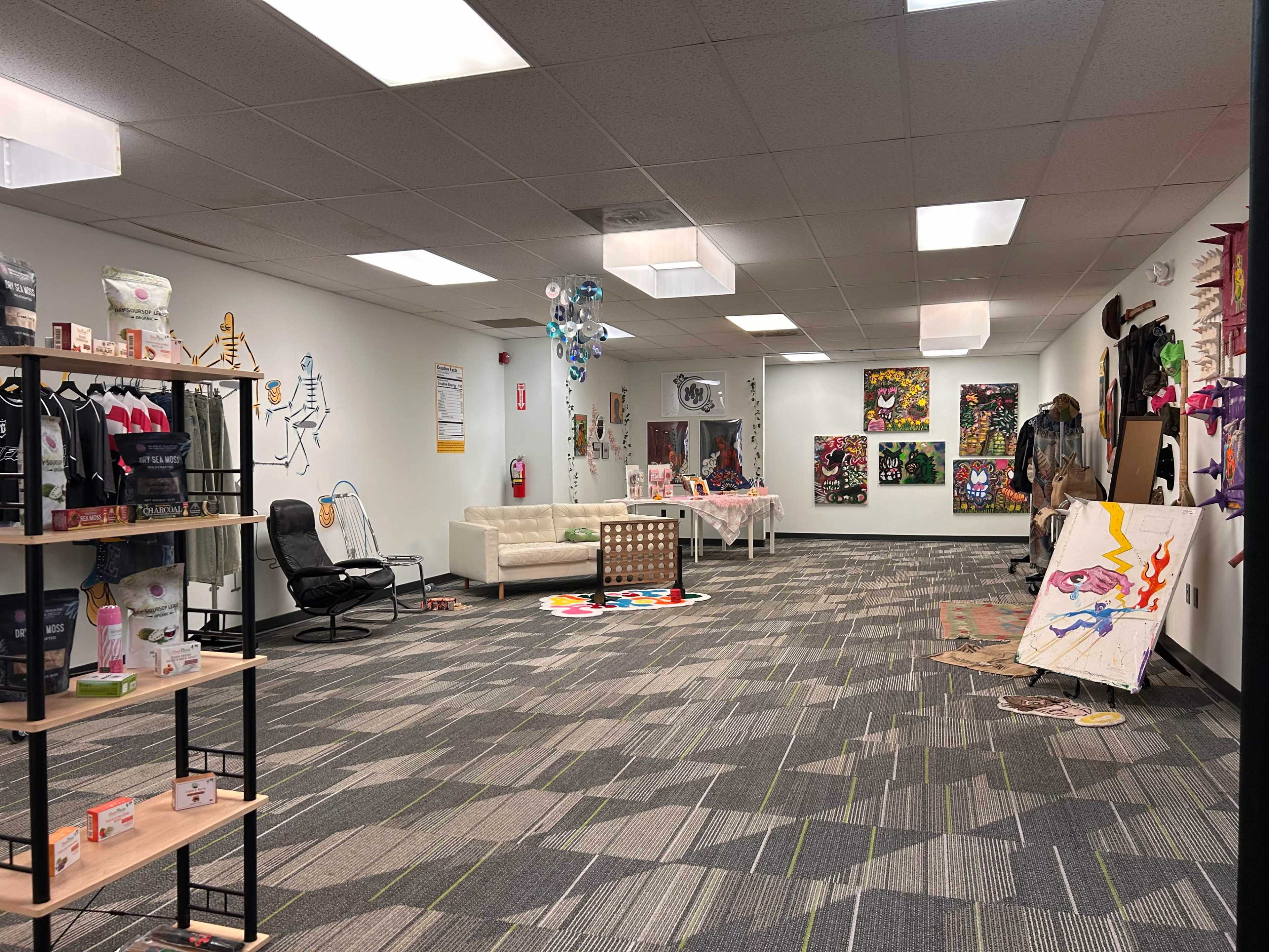 The image shows a spacious room with various artworks displayed on the walls, a white couch, a display shelf with merchandise, and colorful carpeting.