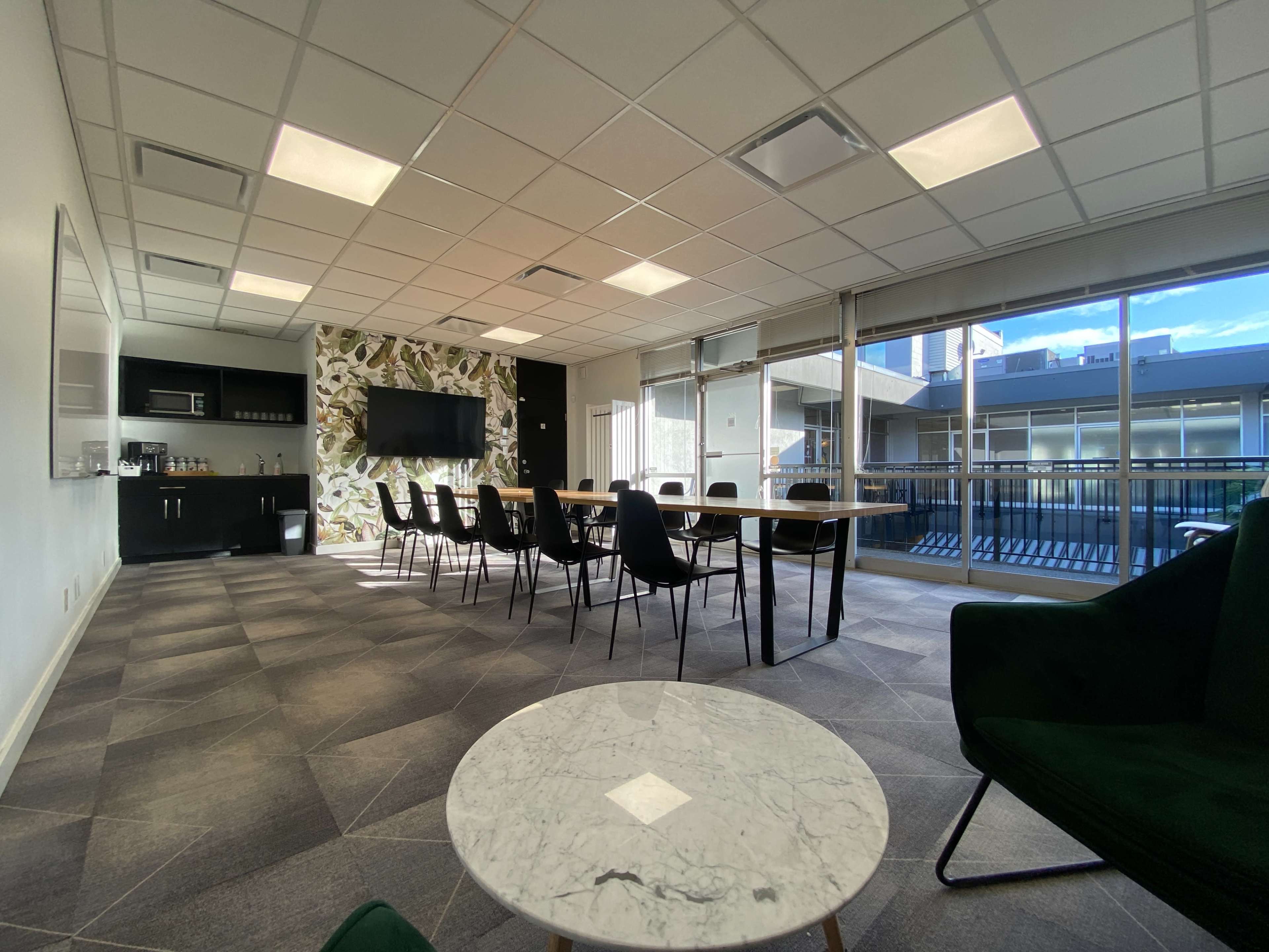 A modern meeting room with a long table, black chairs, and large windows allowing natural light, alongside green foliage decor on one wall.