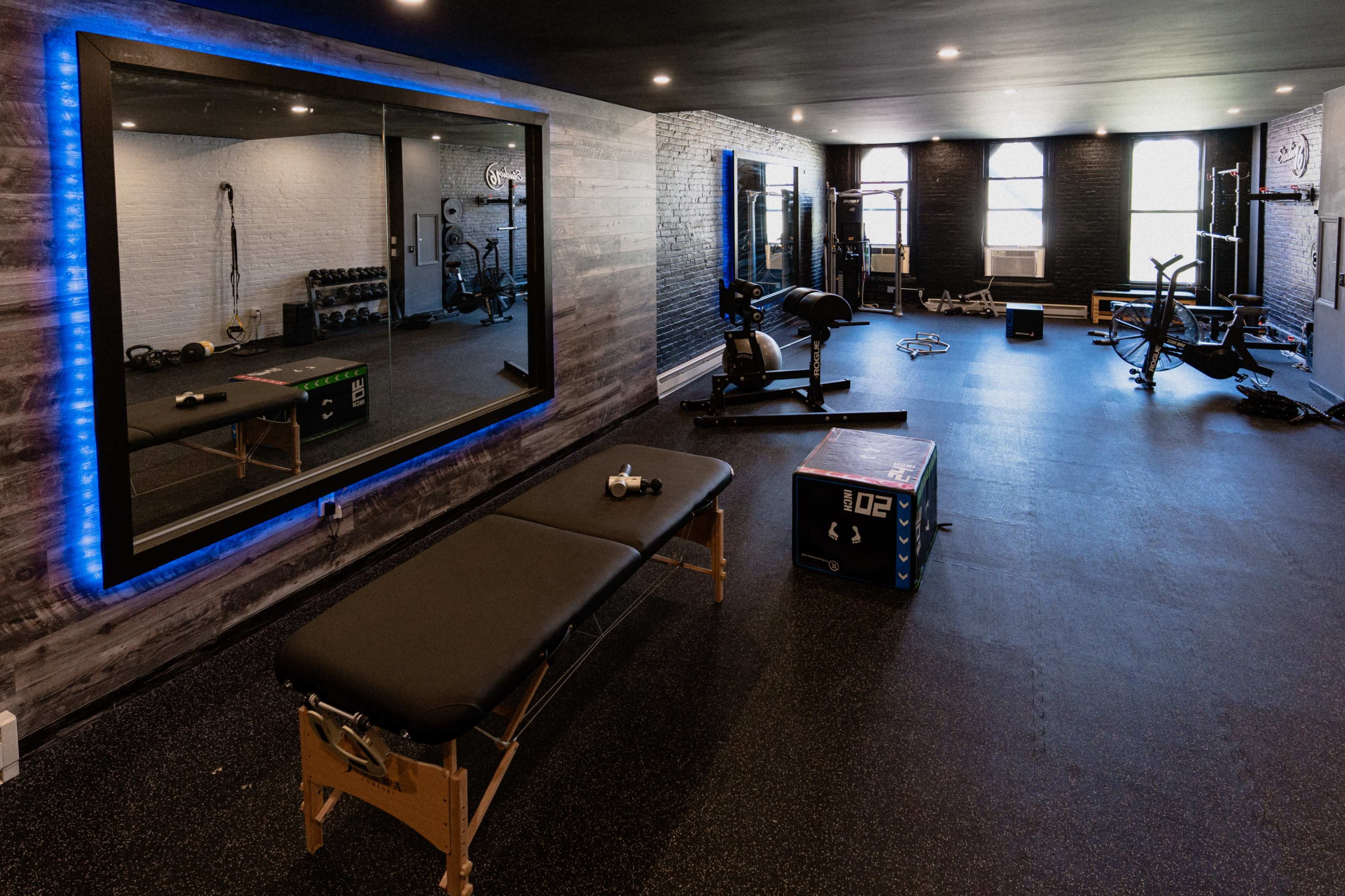 A gym interior with exercise equipment, including a weight bench, dumbbells, and a mirror framed with LED lights.