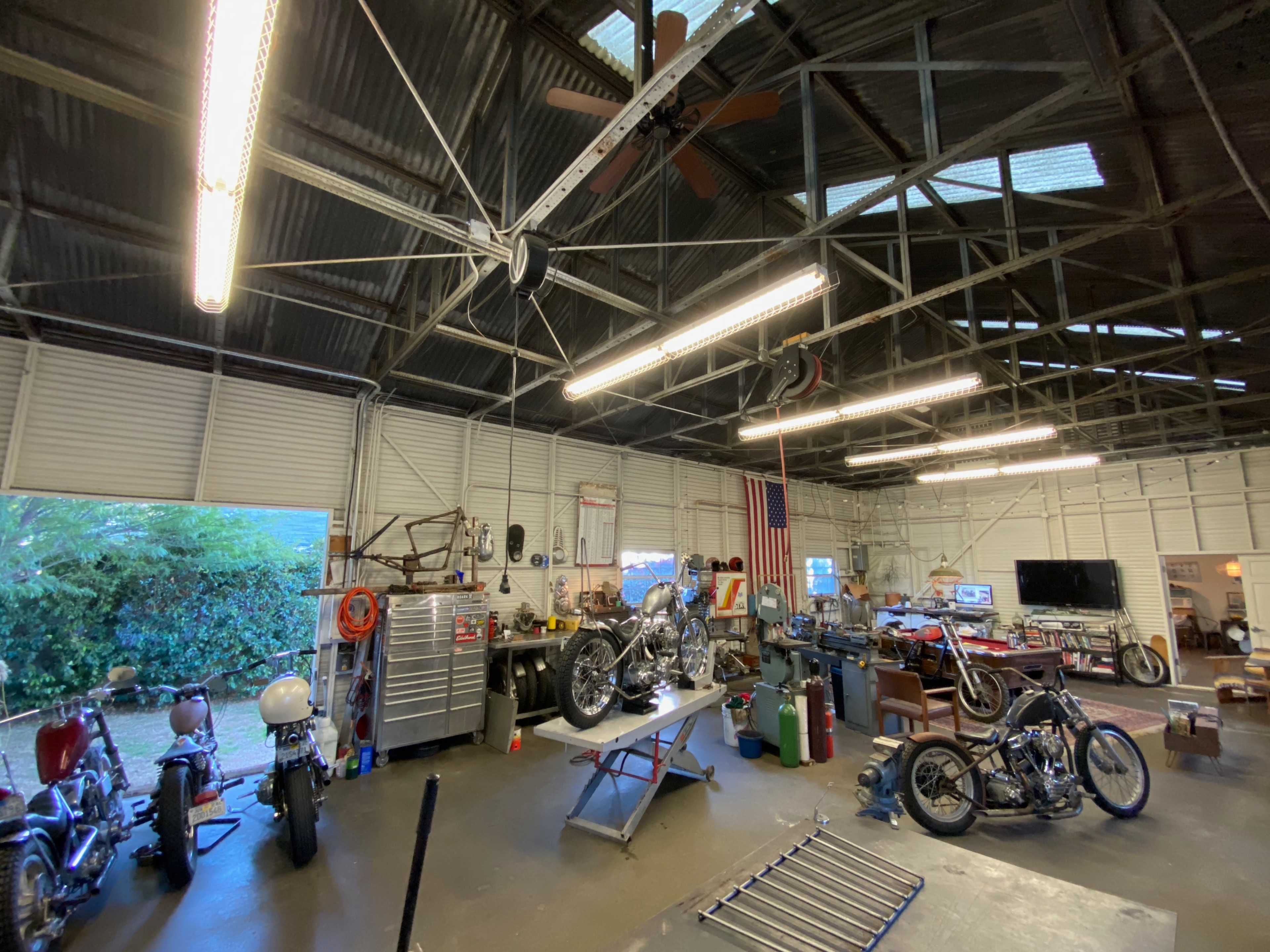 A workshop with multiple motorcycles and tools is illuminated by fluorescent lights under a high metal roof.