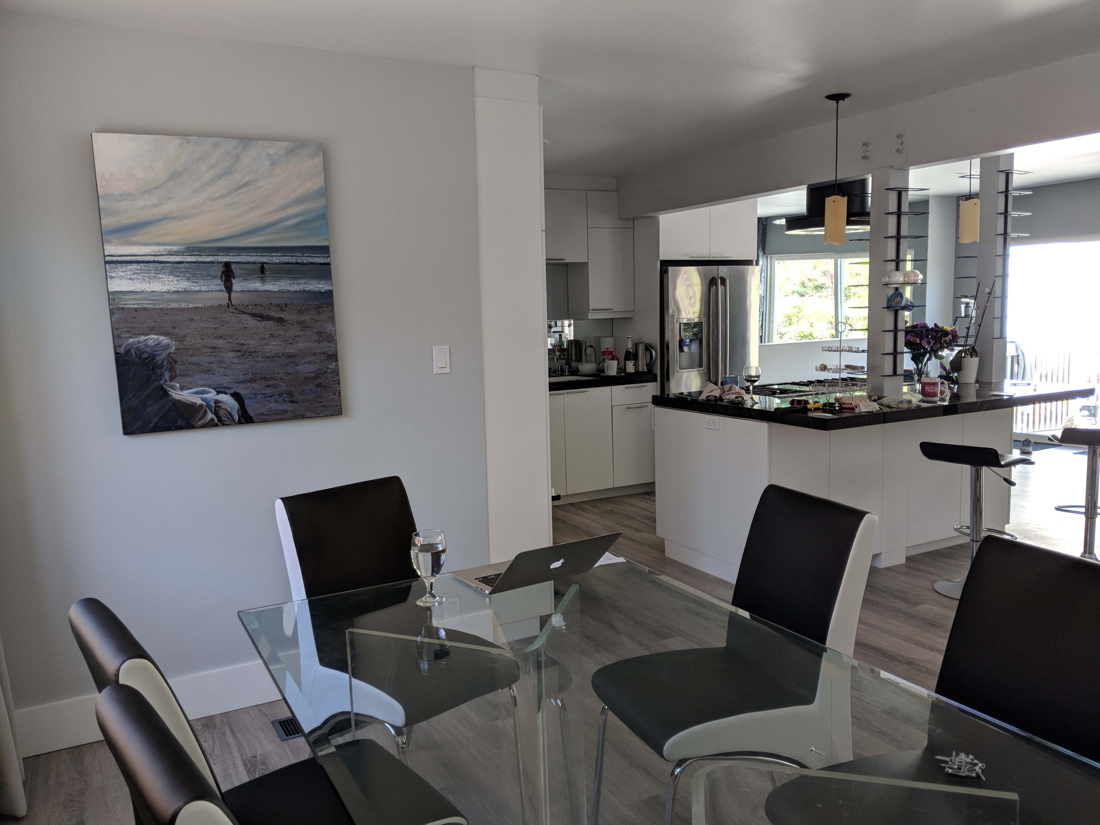 A modern kitchen and dining area features a glass table, black chairs, and a large painting of a beach scene on the wall.