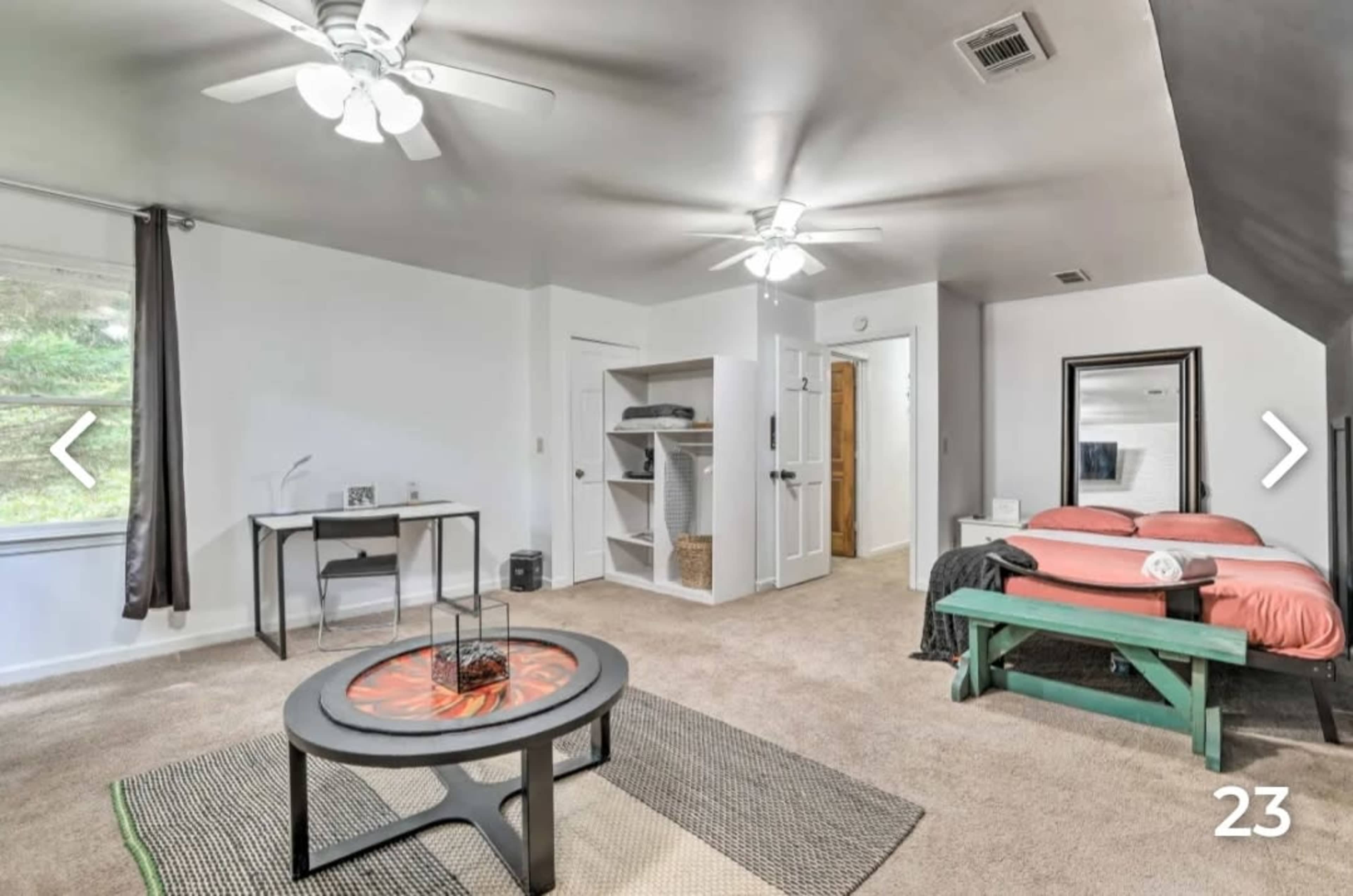 A spacious room features two ceiling fans, a desk, a bookshelf, and a bed with pink bedding, situated near a large window.