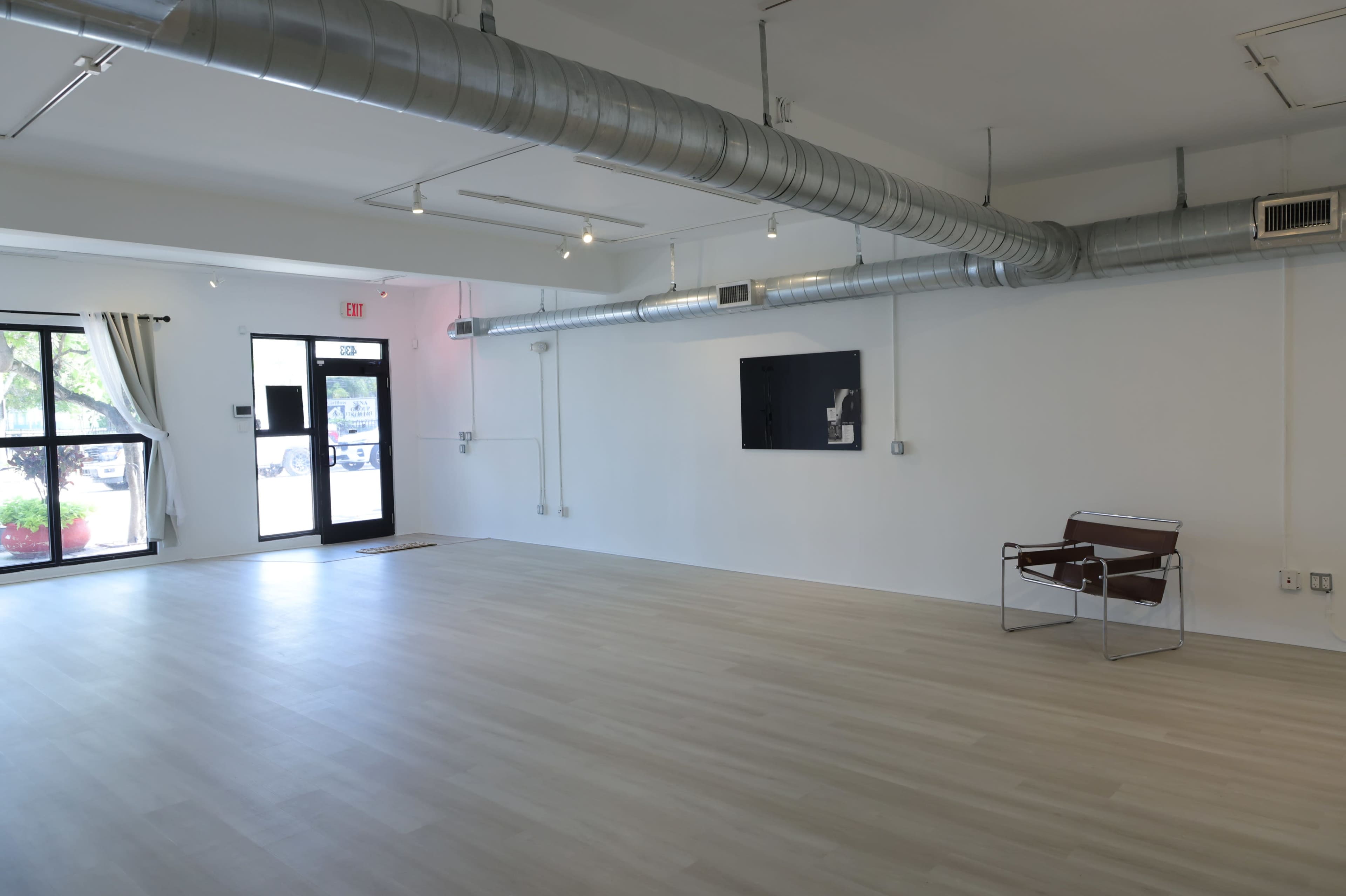 The image shows a spacious, empty room with light-colored flooring, exposed metal ductwork on the ceiling, and a minimalist chair positioned in one corner.