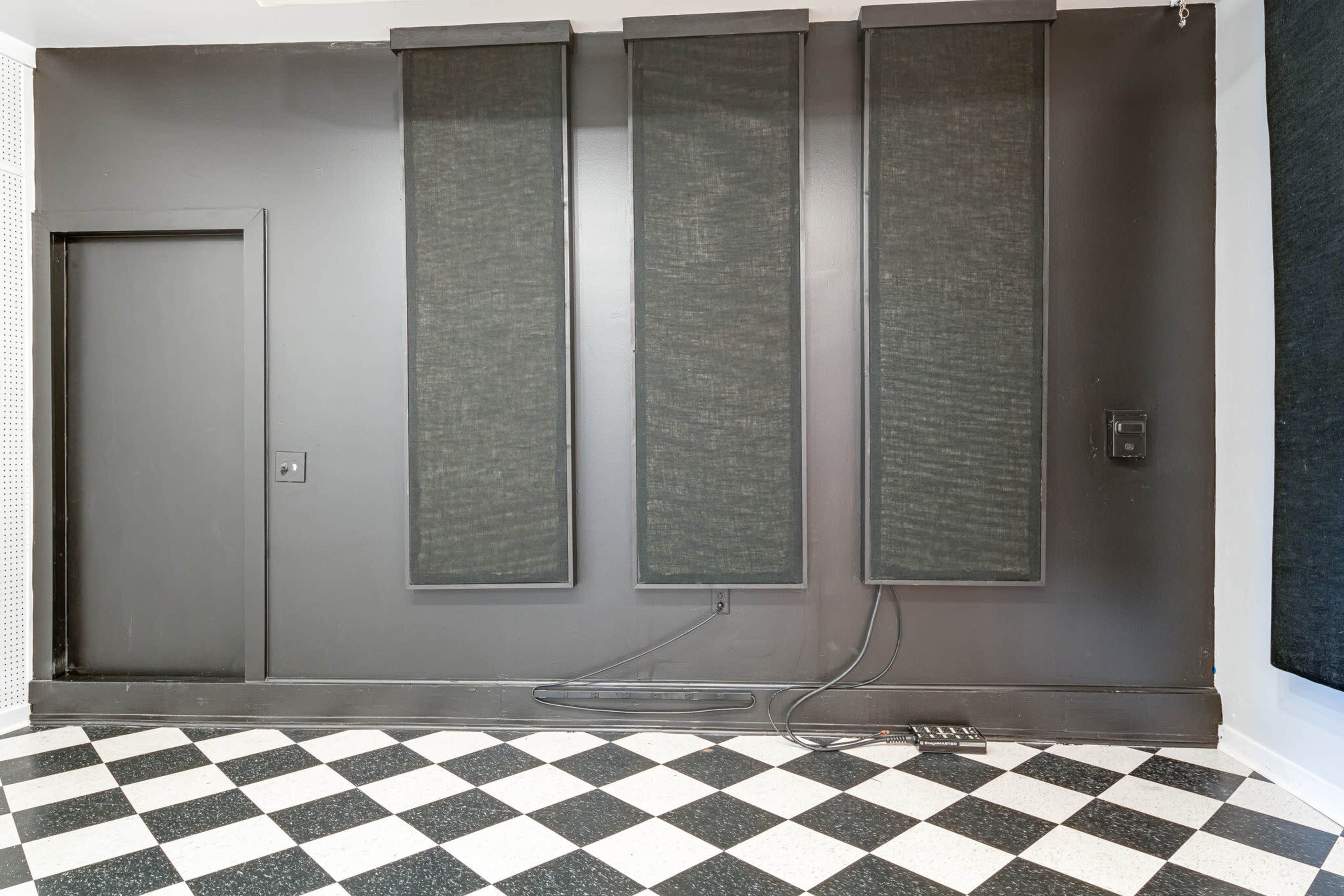 A room with a black wall featuring three large sound-absorbing panels and a door, alongside a checkered black and white floor.