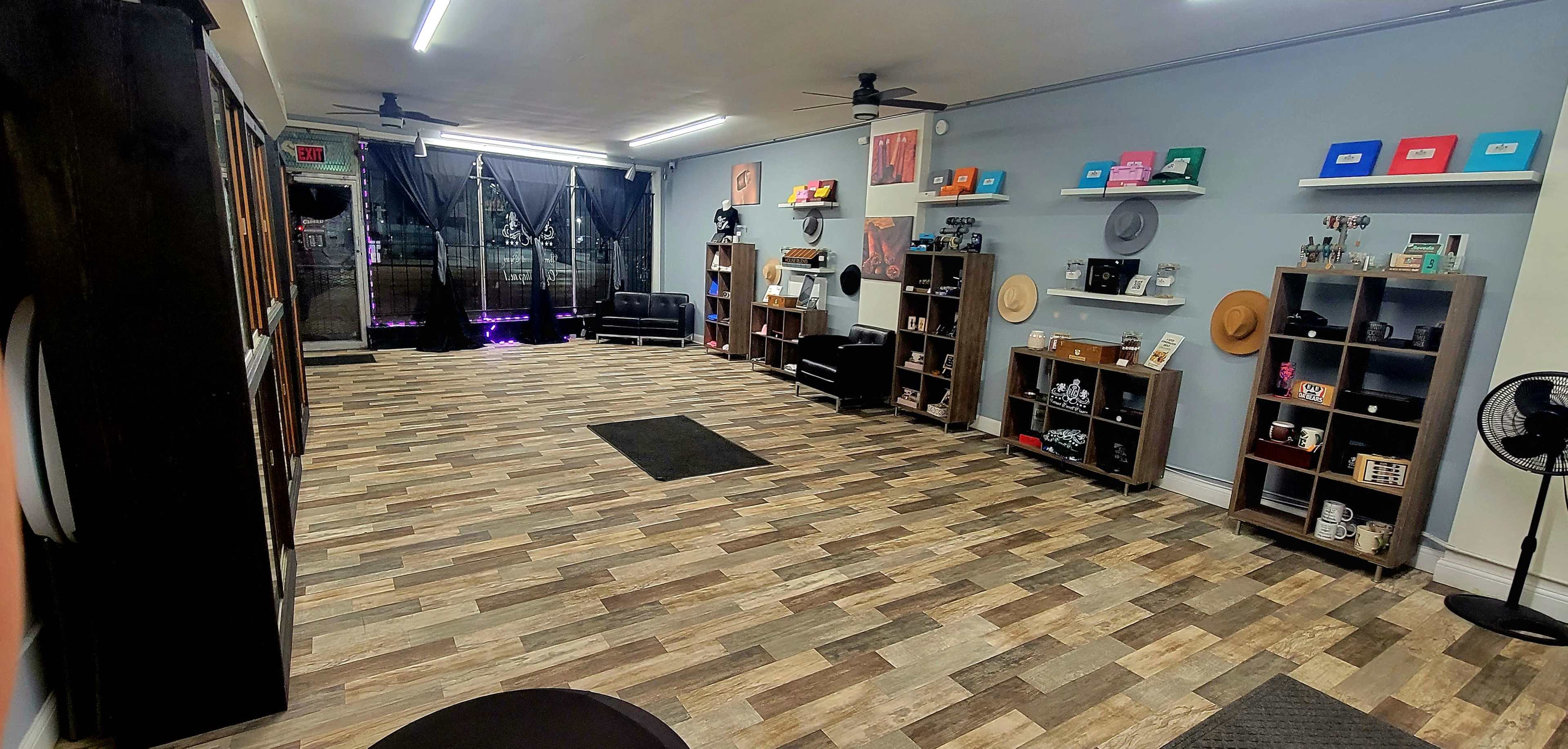 The image shows a spacious indoor area with wooden flooring, featuring shelves displaying various items, two seating areas with black couches, and a large window with black curtains.