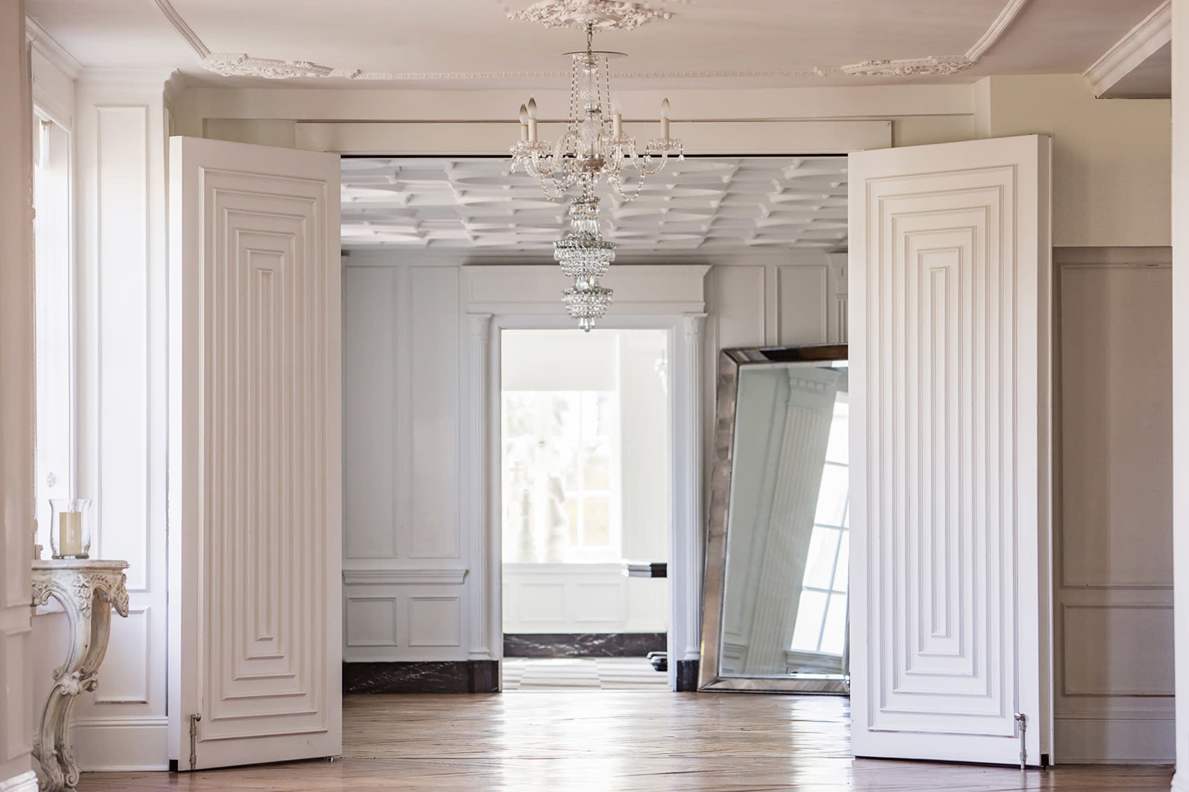 The image features an elegantly designed interior space with white doors open to reveal a bright room that includes a large mirror and a chandelier.