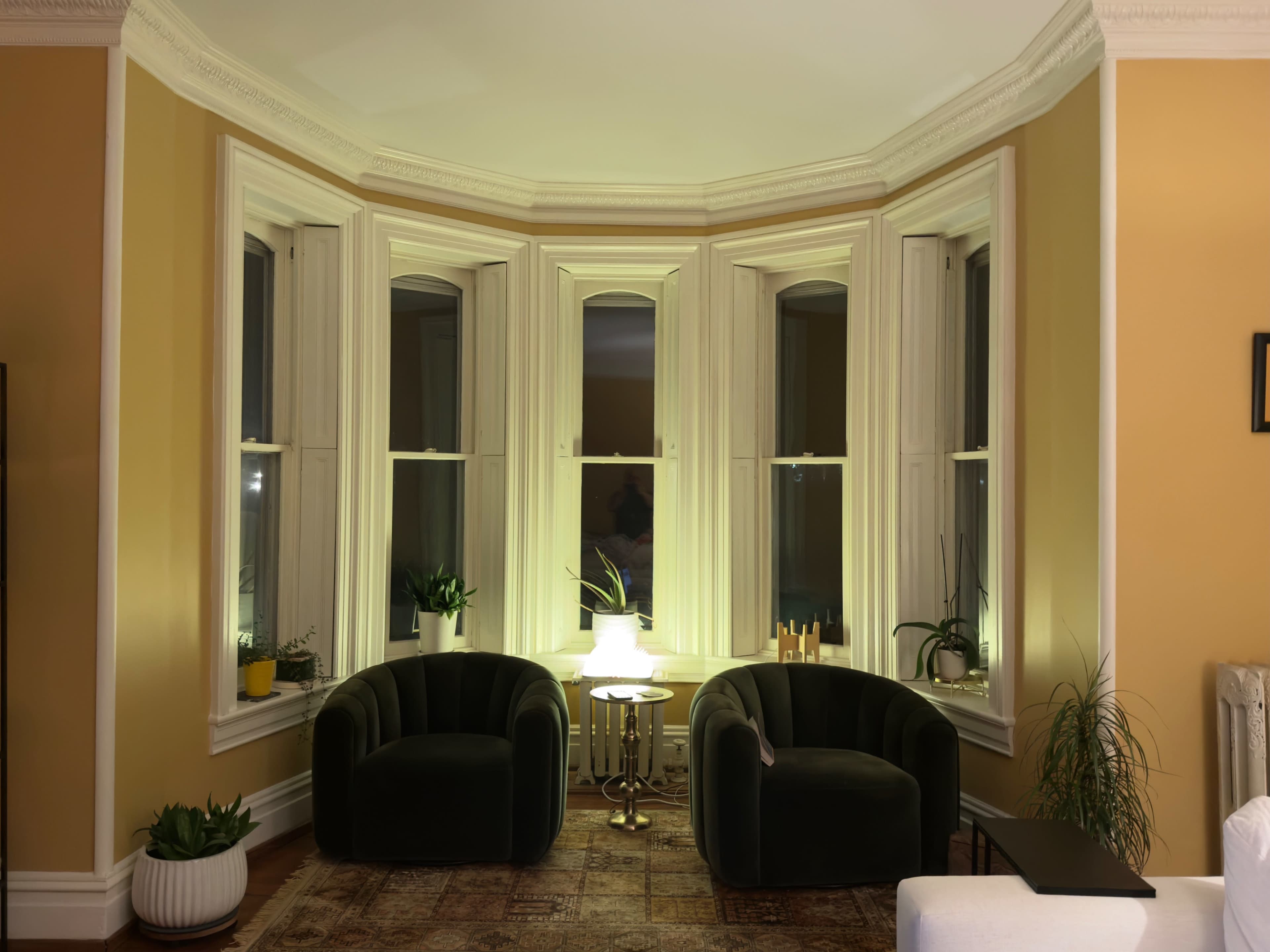 A cozy corner of a room features two black armchairs beside a small table, illuminated by warm lighting, and surrounded by plants and large windows.