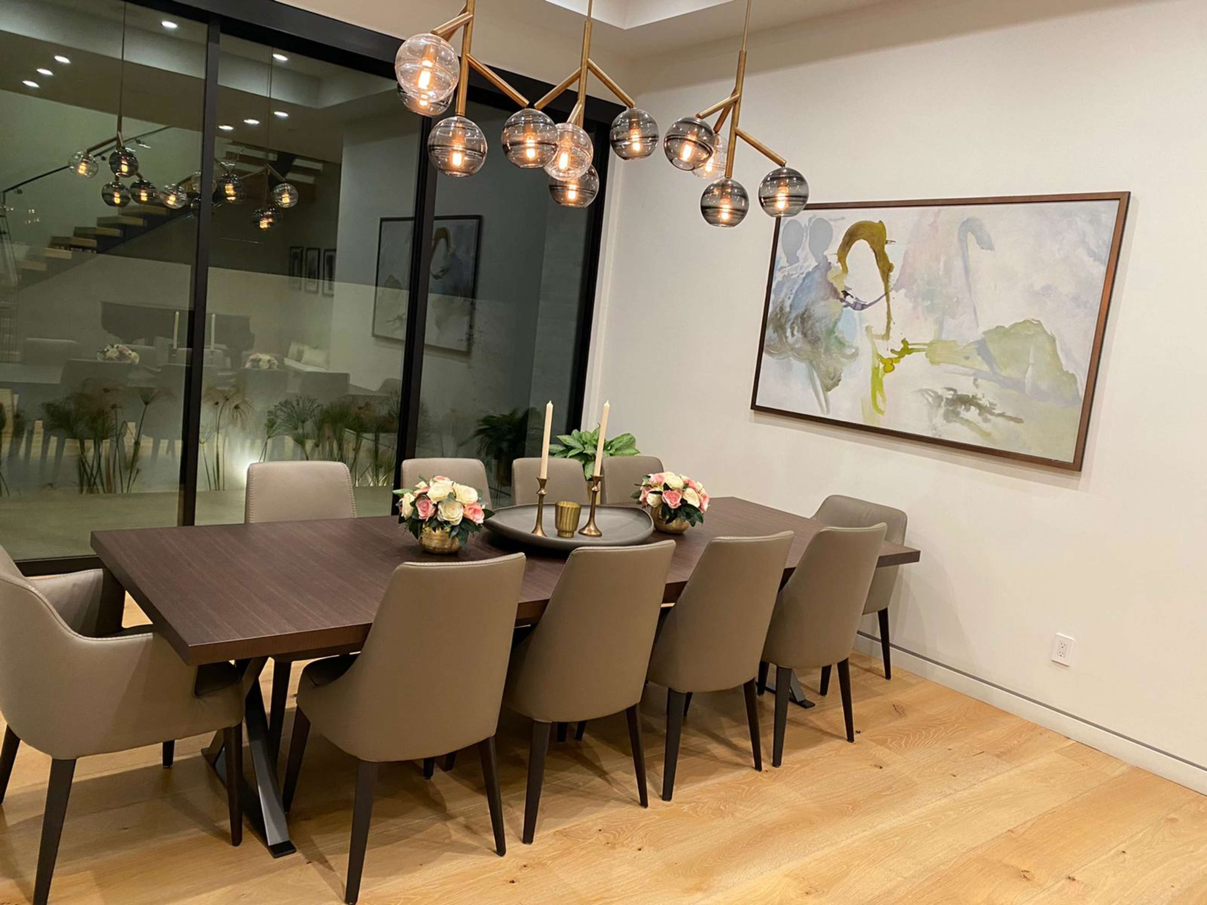 A modern dining room features a large wooden table surrounded by upholstered chairs and a statement light fixture above, with a large window showcasing a view of the exterior.