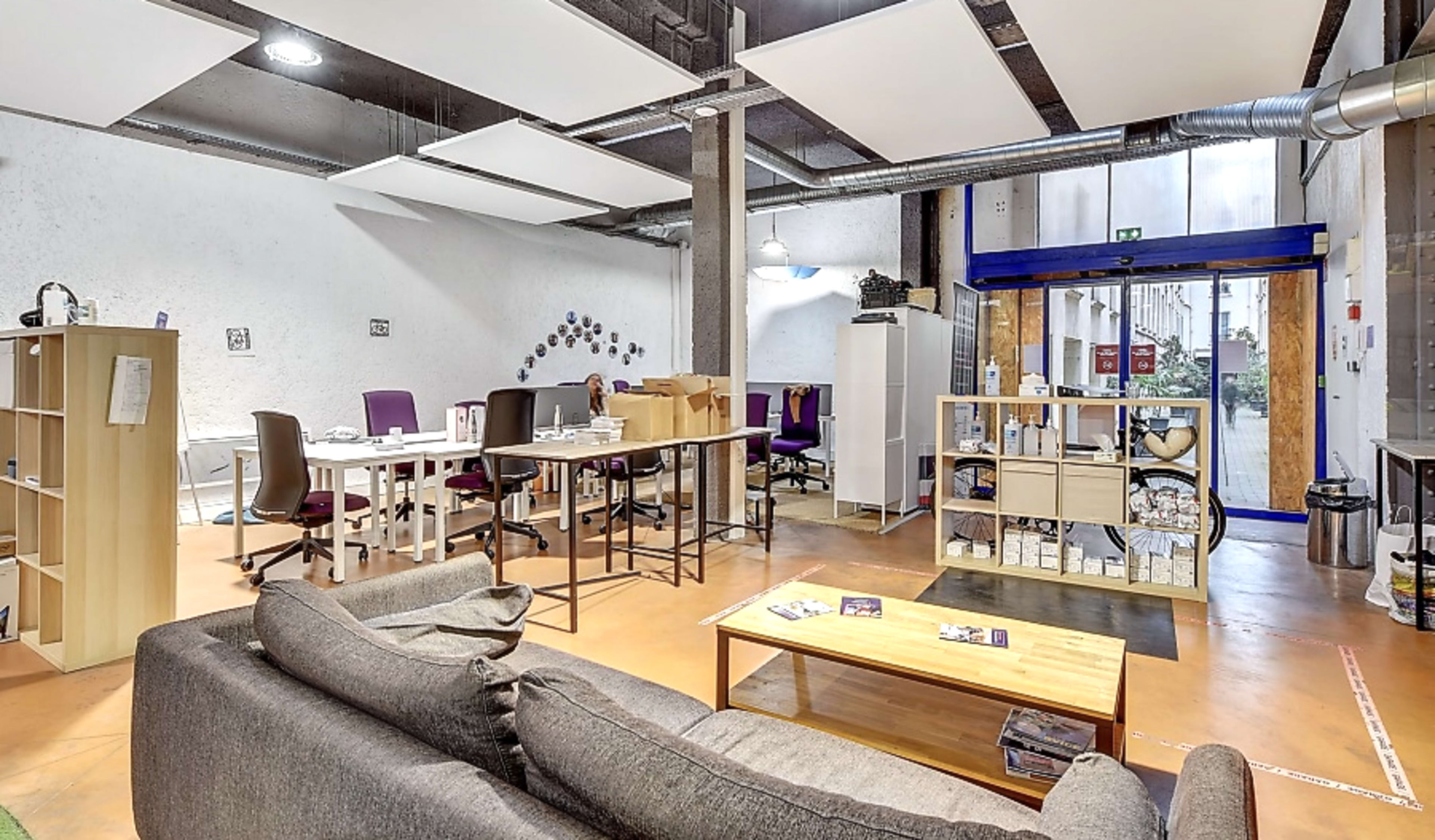 The image shows a modern office space with a seating area, workstations, and organized shelving units.