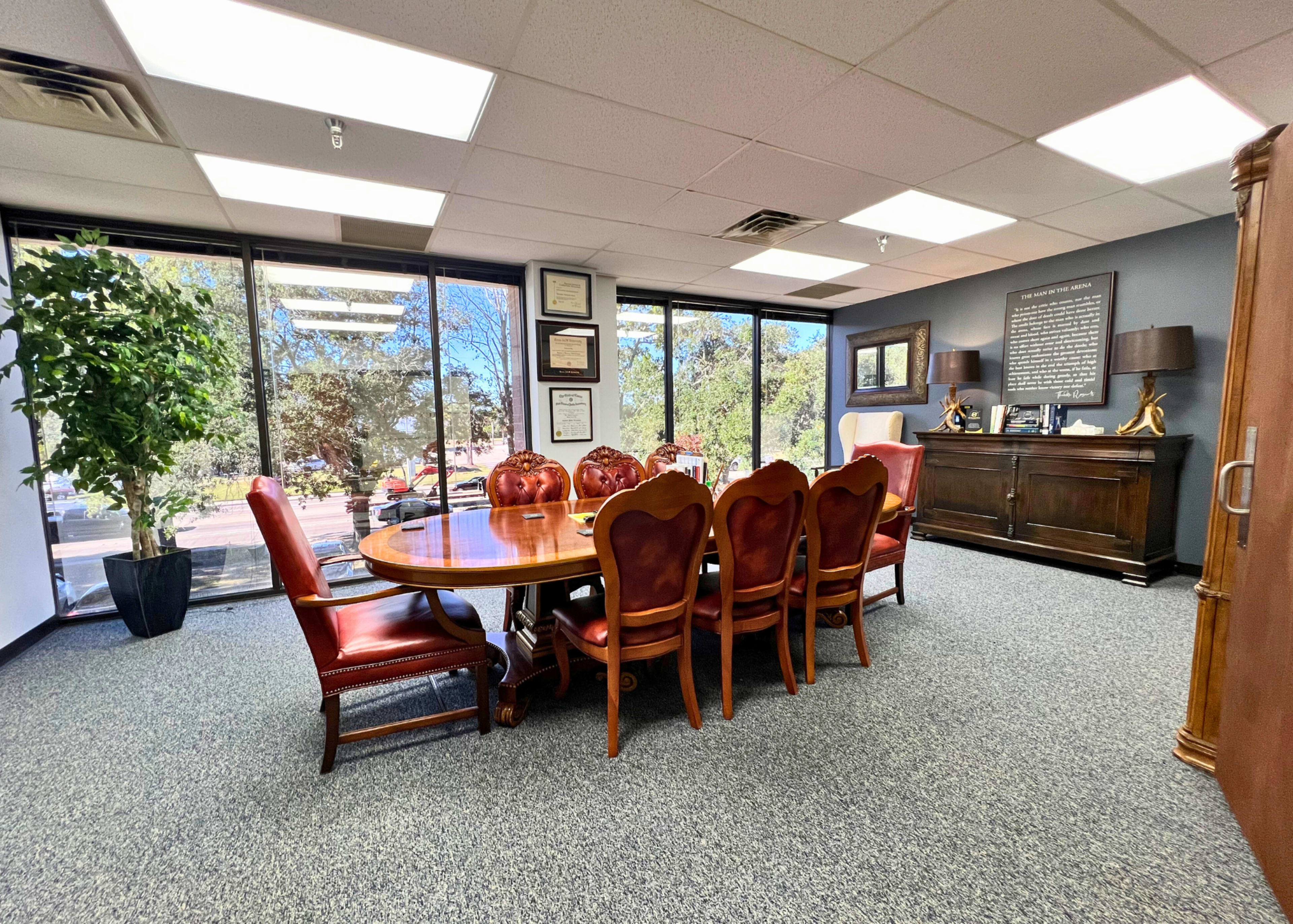 A conference room featuring a large wooden table surrounded by eight red upholstered chairs, with large windows providing natural light and a view of greenery outside.