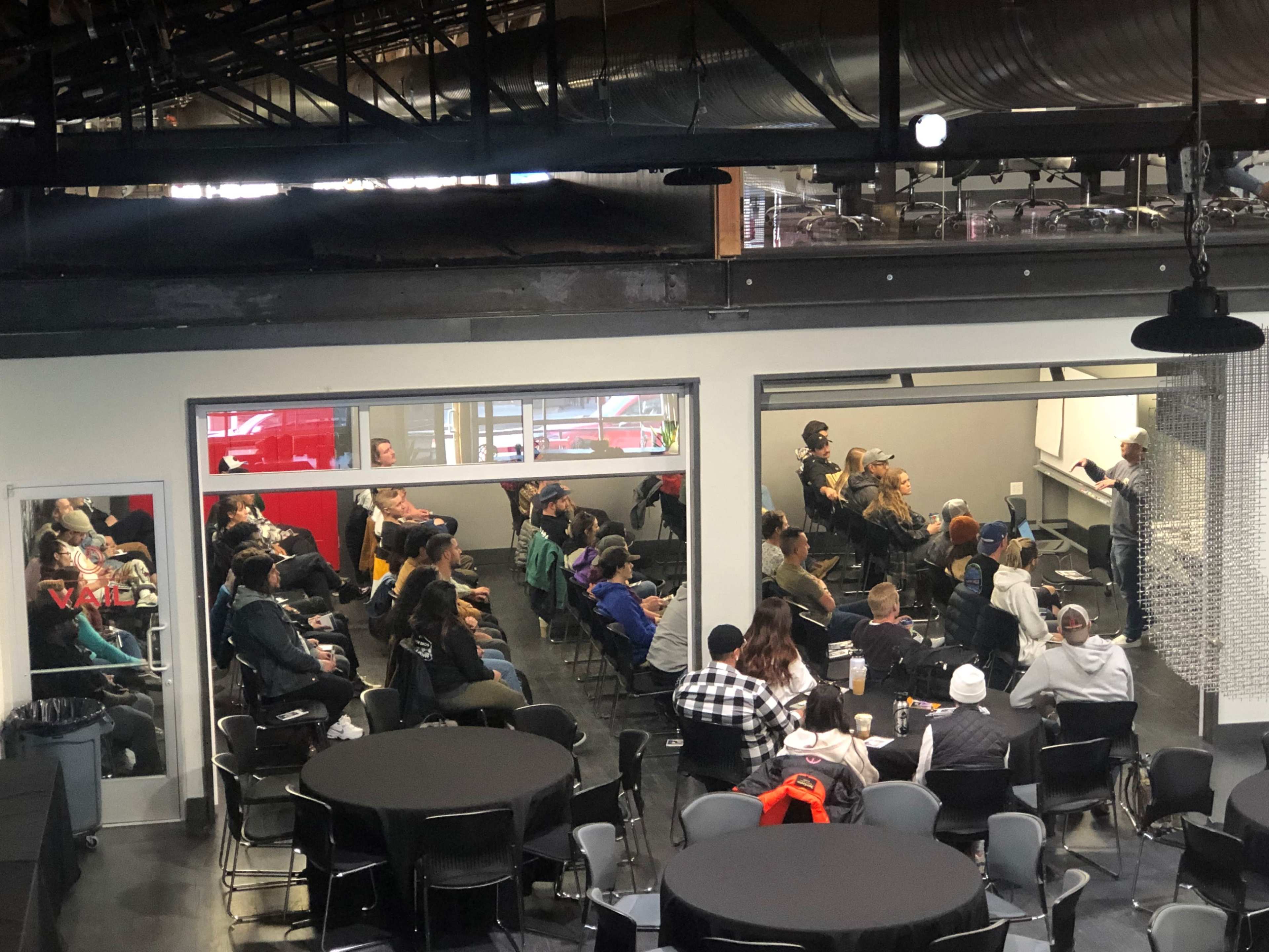 A group of people sits in chairs facing a speaker in a seminar room, while tables are set up in a nearby area.