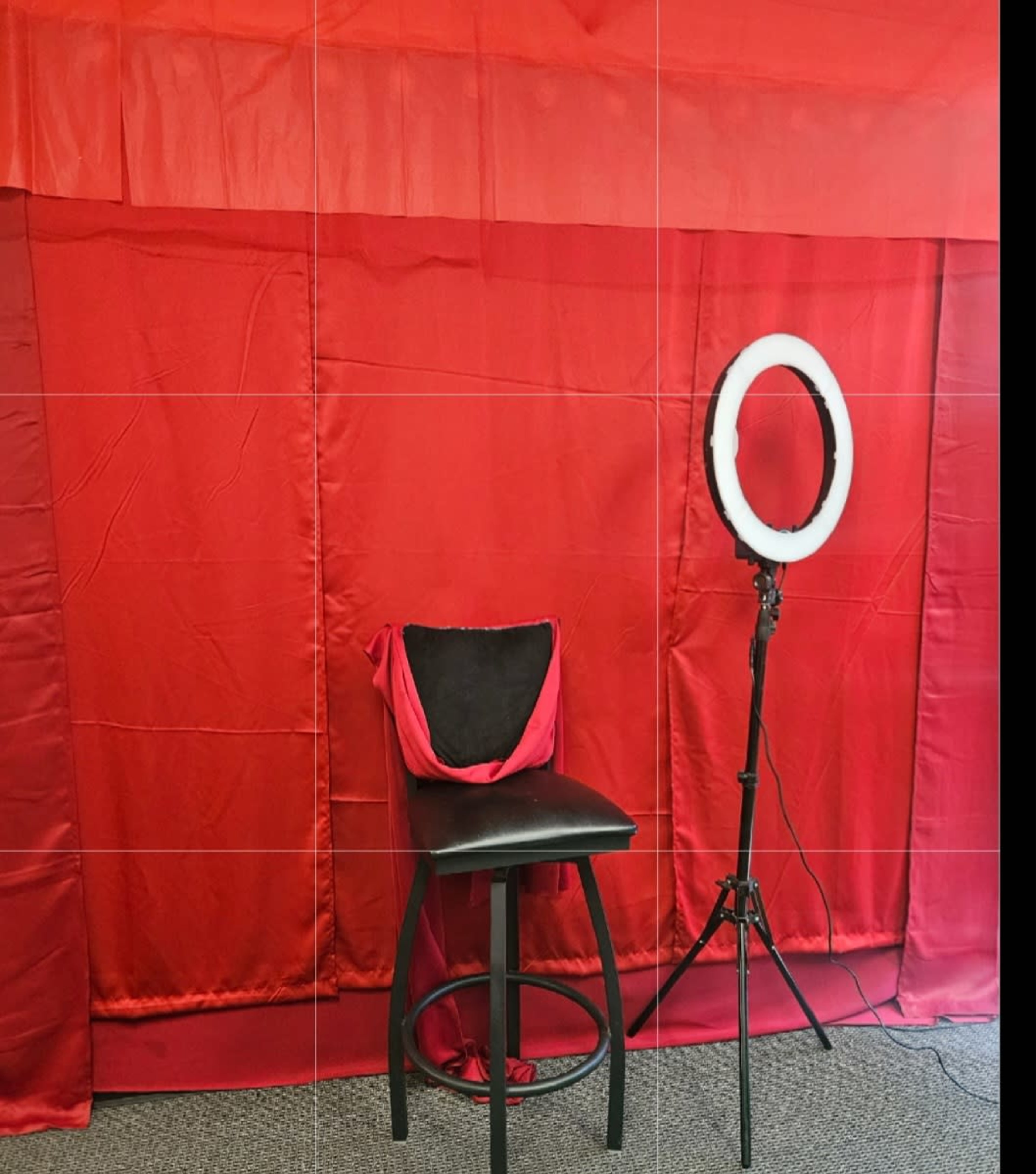 The image shows a black stool with a red and black pillow in front of a red backdrop and a ring light on a stand.