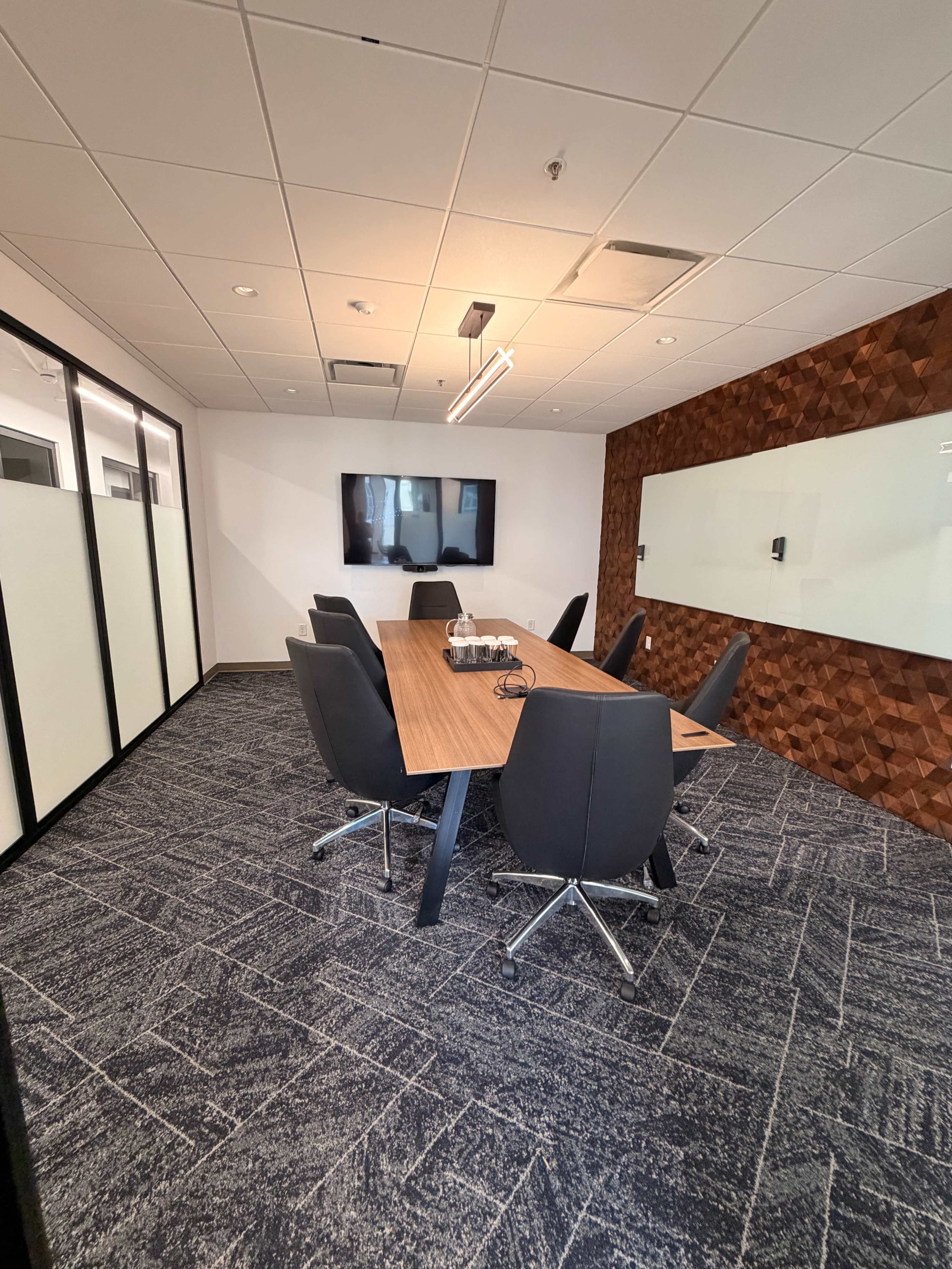 The image shows a modern conference room with a long wooden table, six black chairs, a wall-mounted monitor, and a textured wood accent wall.