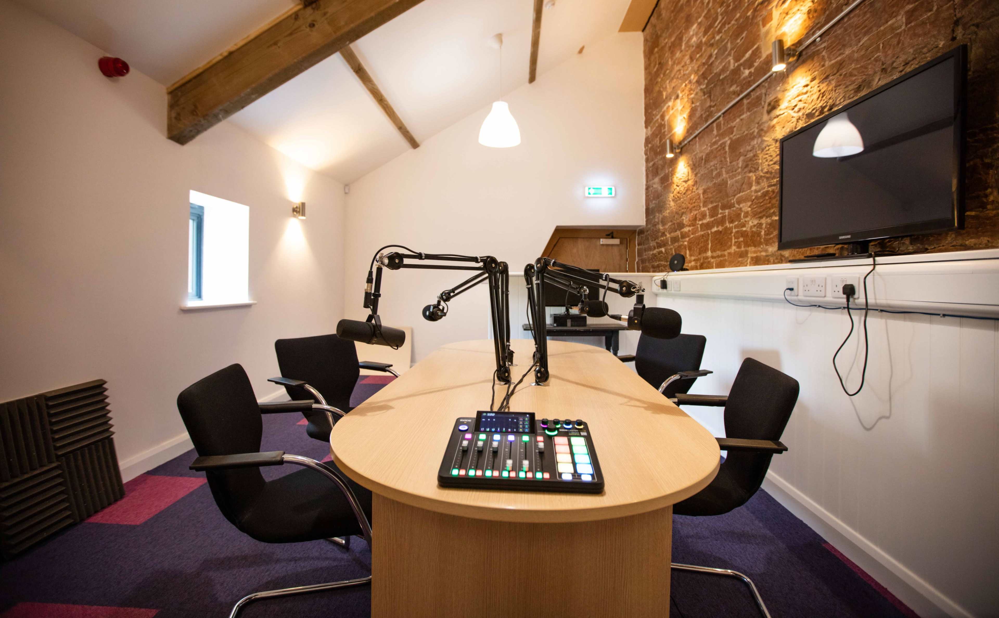 The image shows a podcast recording studio with a long table, four black chairs, and a wall-mounted TV, featuring audio equipment in the center.