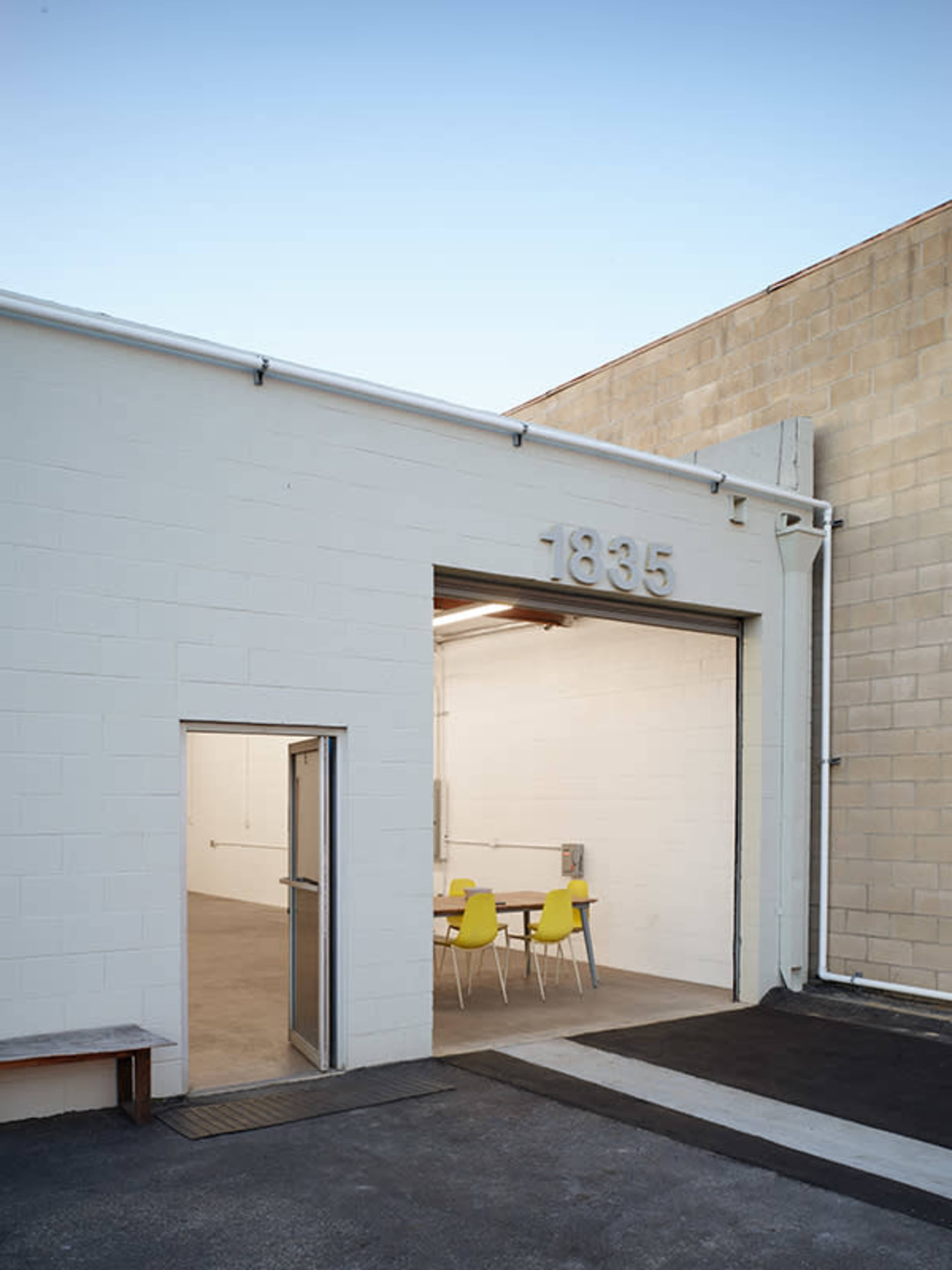 A simple, white warehouse exterior with the number 1835 displayed above an open entrance leading into a sparsely furnished interior with yellow chairs around a table.