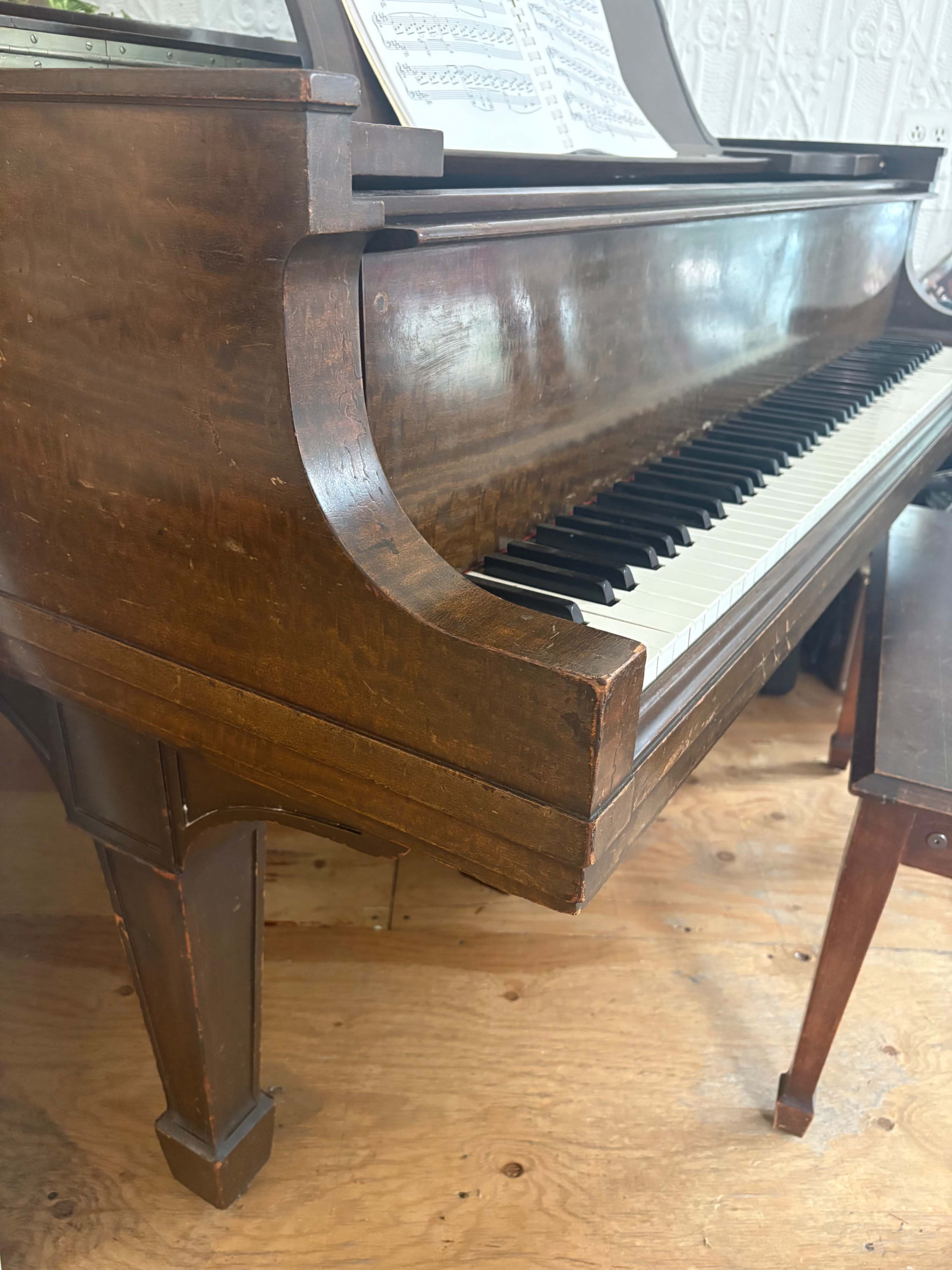 The image shows a wooden piano with a polished surface and black and white keys, positioned next to a small stool on a wooden floor.