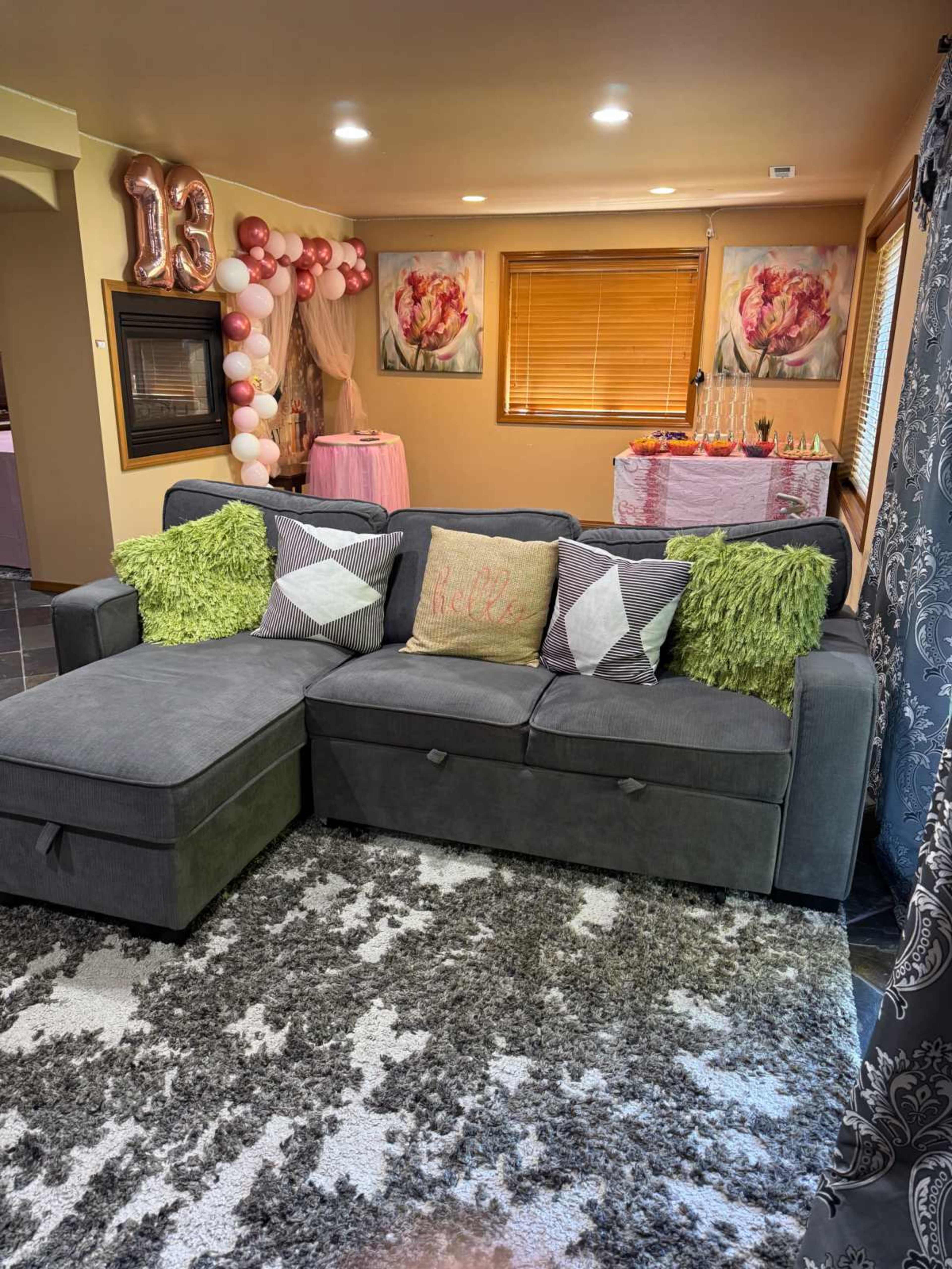 The image shows a living room with a gray sectional sofa adorned with decorative pillows, a large area rug, and a festive setup featuring a table decorated with balloons and flowers in the background.