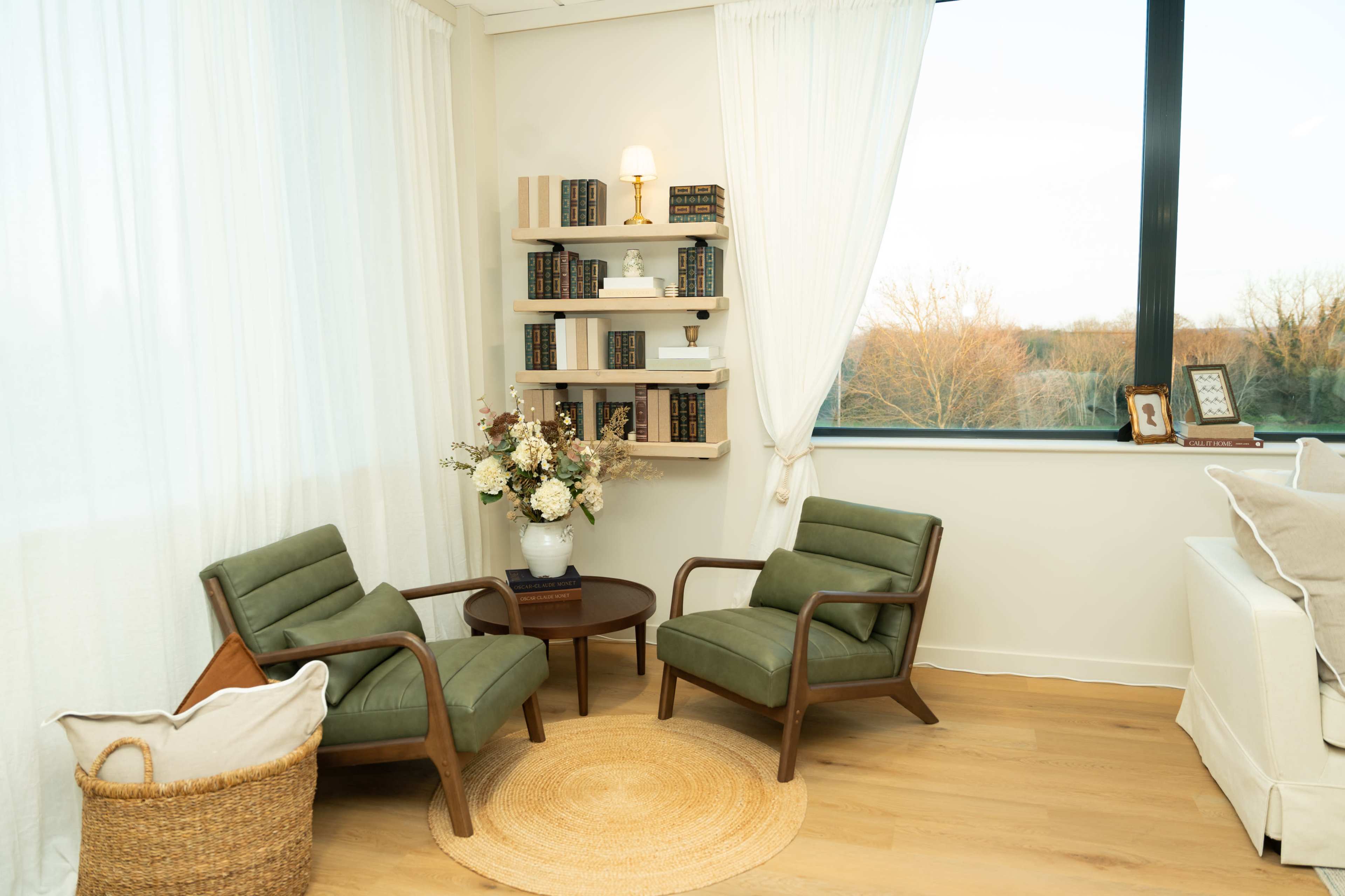 The image depicts a cozy reading nook with two green armchairs, a circular wooden table, and a bookshelf filled with books, alongside a floral arrangement, all situated near a large window.