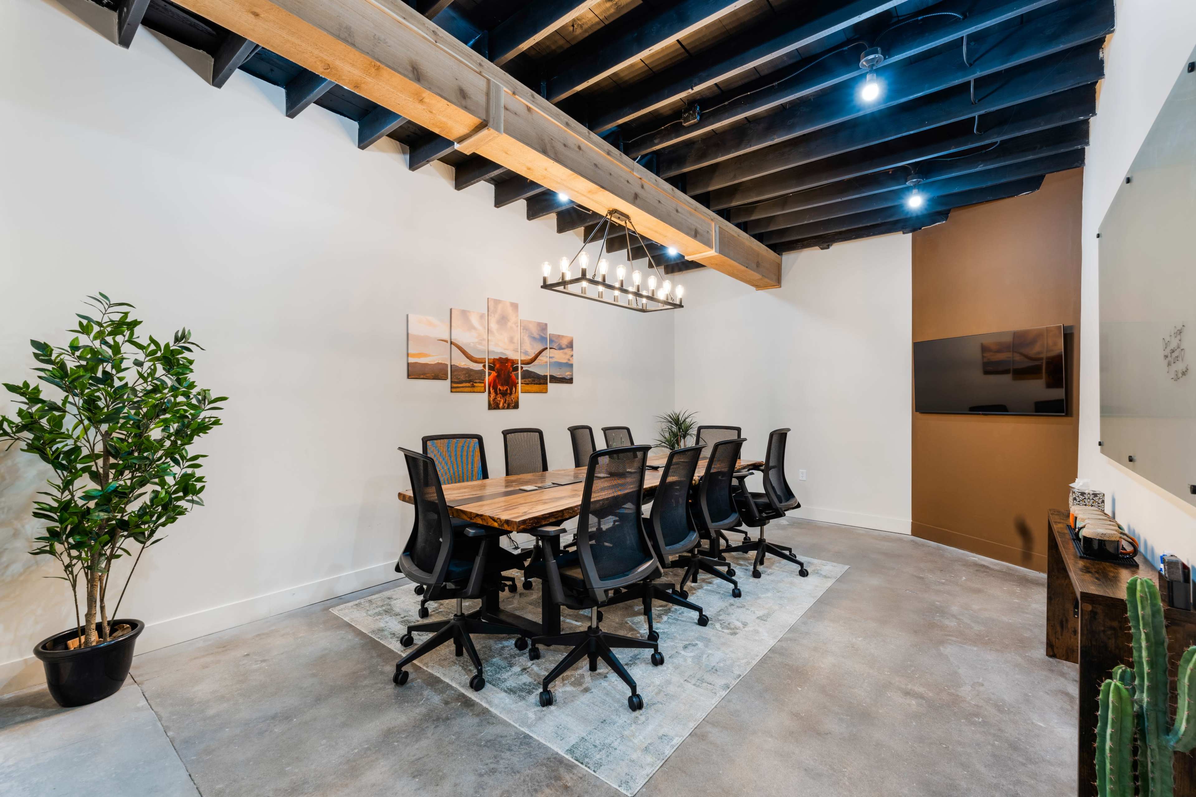 A conference room features a long wooden table surrounded by ergonomic chairs, with a large chandelier and plants decoratively positioned in the space.