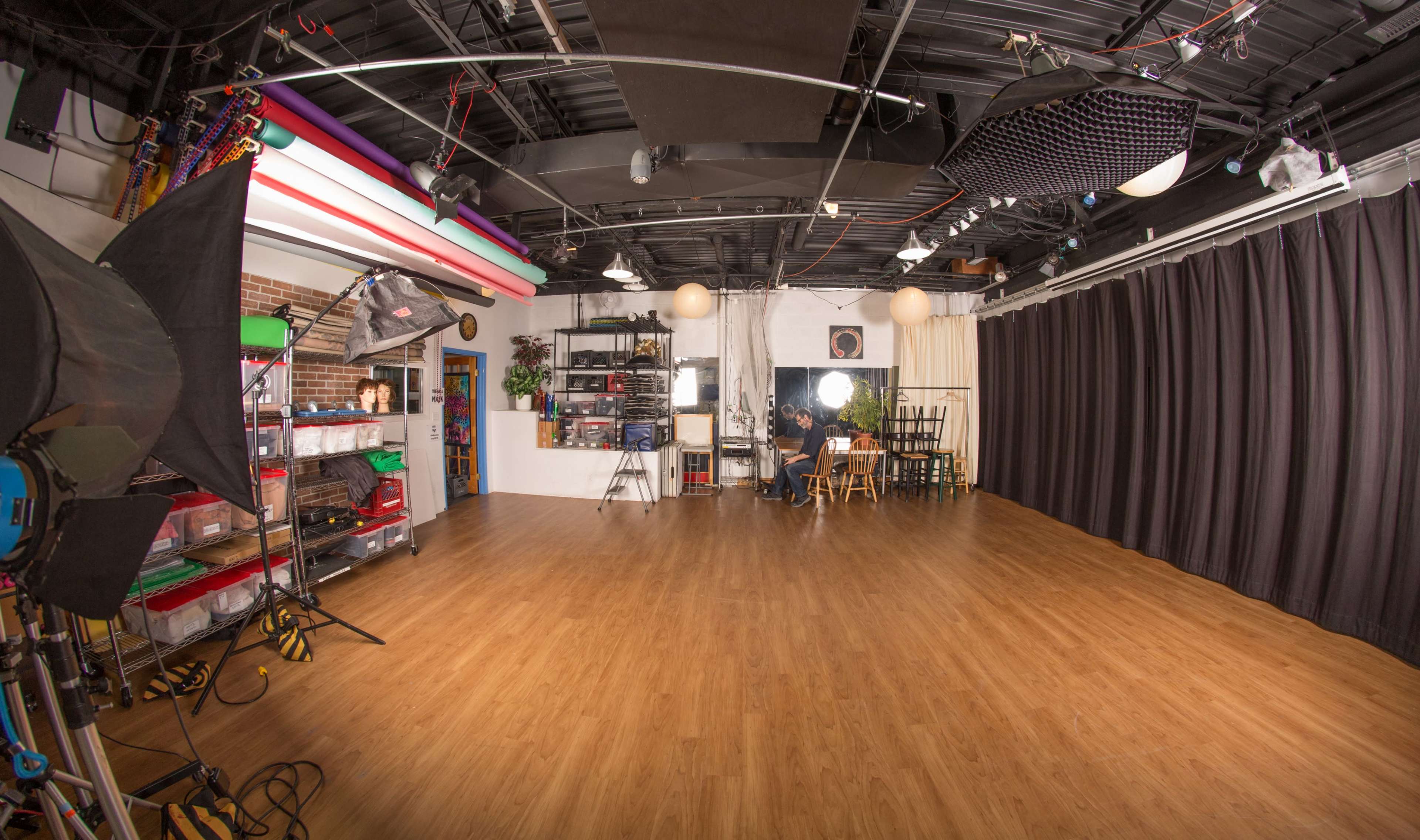 A photography studio with various lighting equipment, a wooden floor, and a backdrop of black curtains.