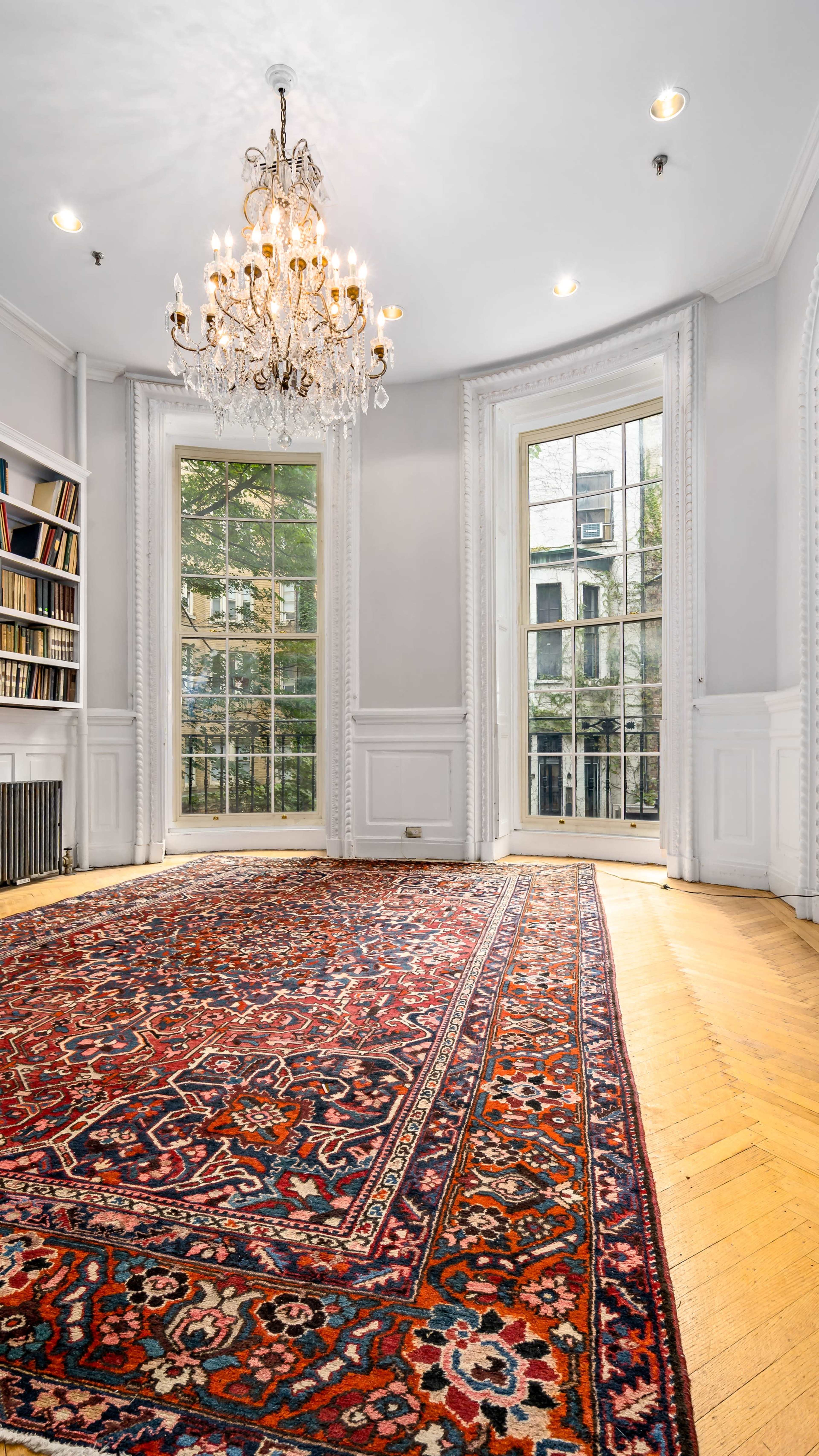 The image shows a well-lit room with large windows, a chandelier, a bookshelf along one wall, and a decorative rug on the wooden floor.