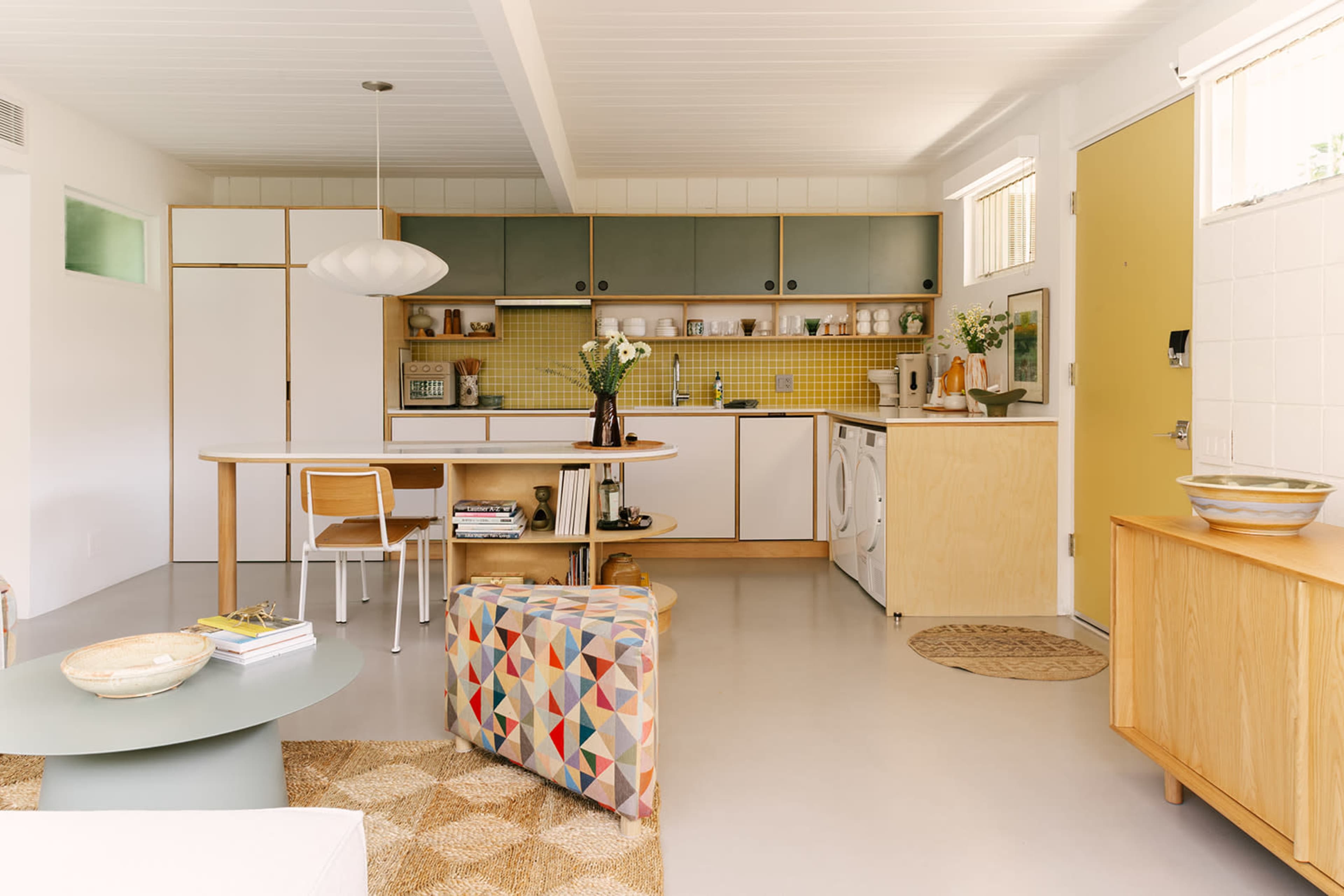 The image shows a modern kitchen with a circular wooden dining table, colorful cabinetry, a yellow door, and appliances arranged in an open layout.