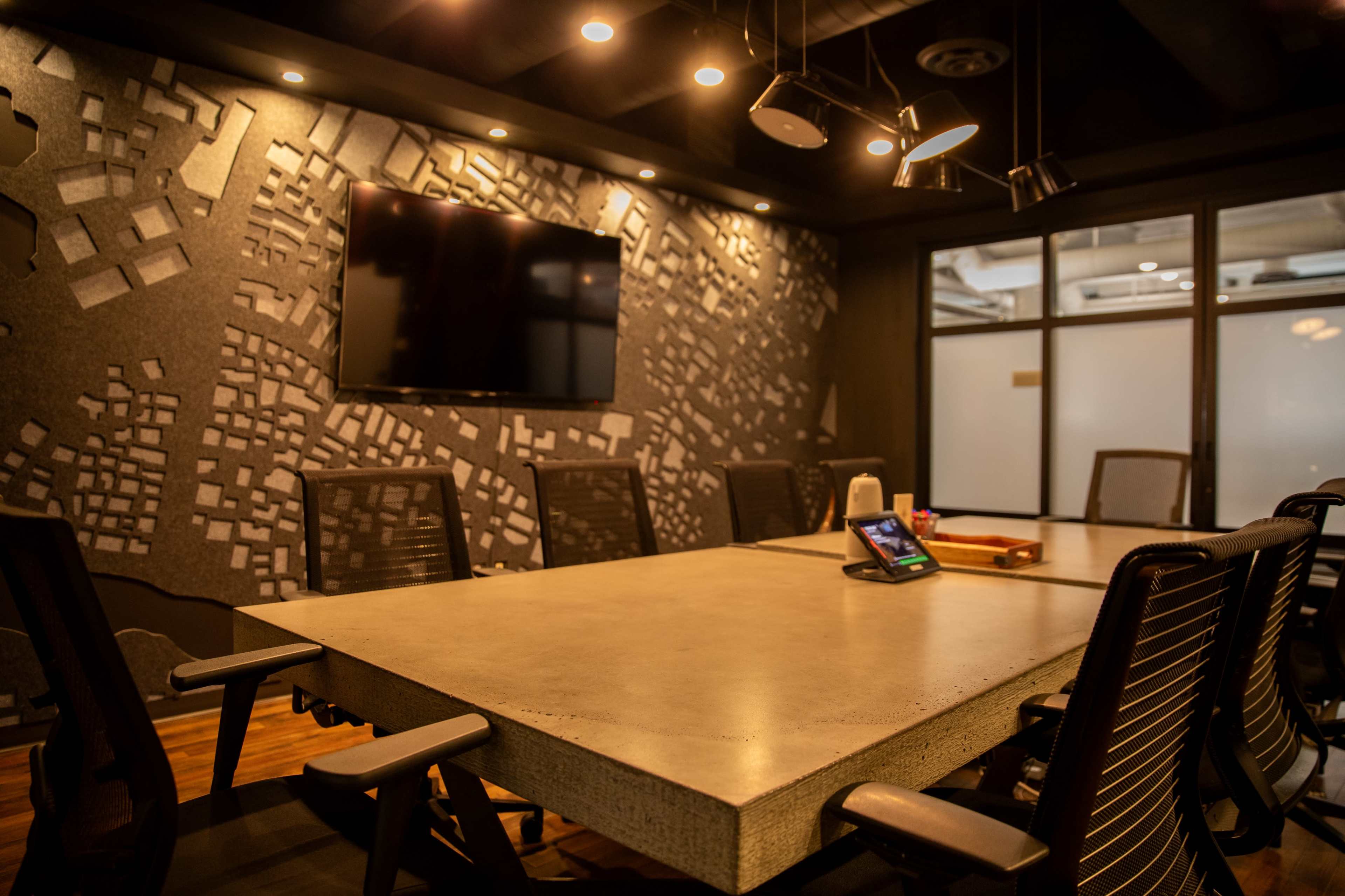 The image depicts a modern conference room with a large concrete table, black mesh chairs, a wall-mounted screen, and a textured design on the wall.