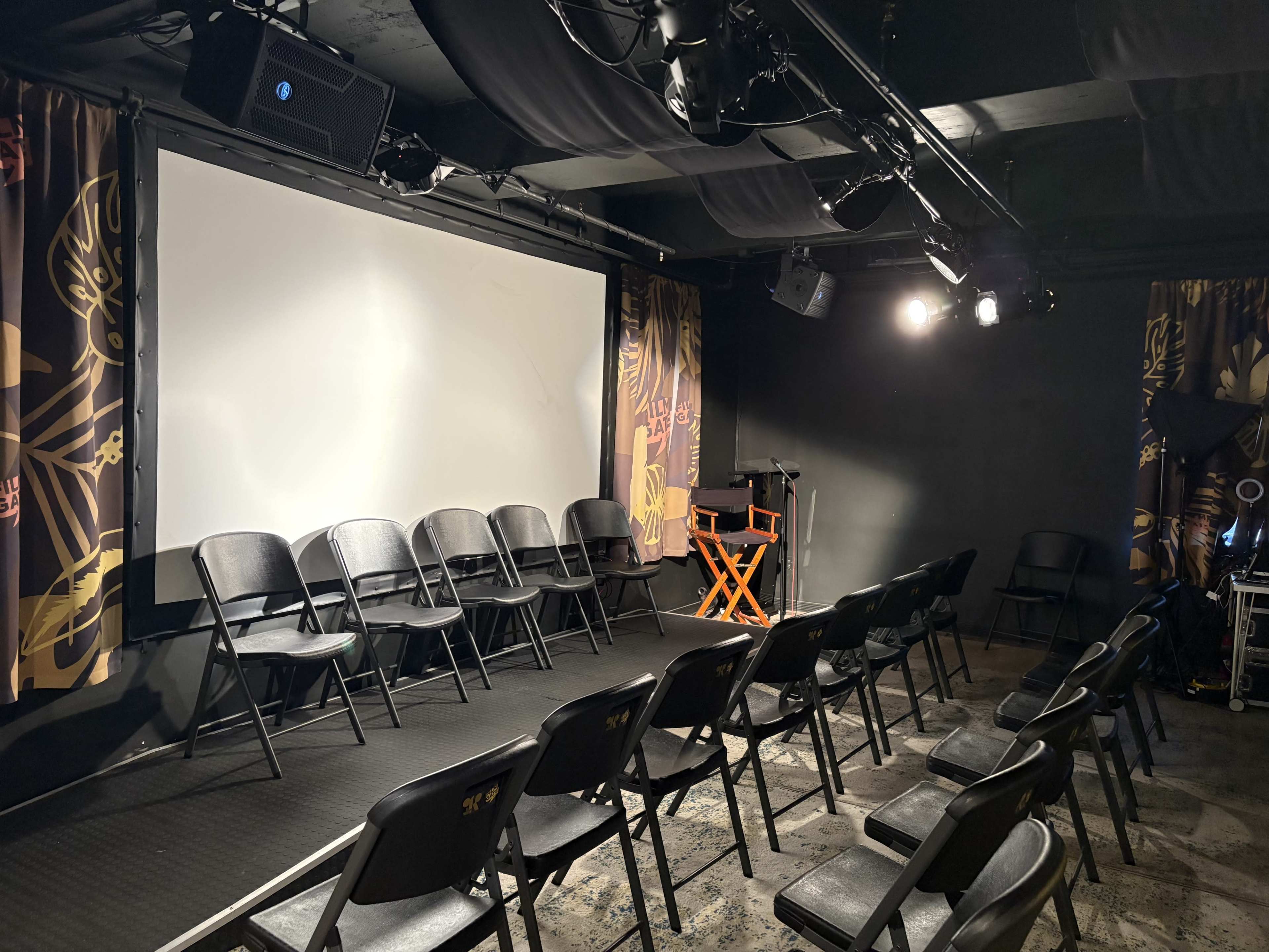 The image shows a small theater setup with rows of black chairs facing a large white screen.