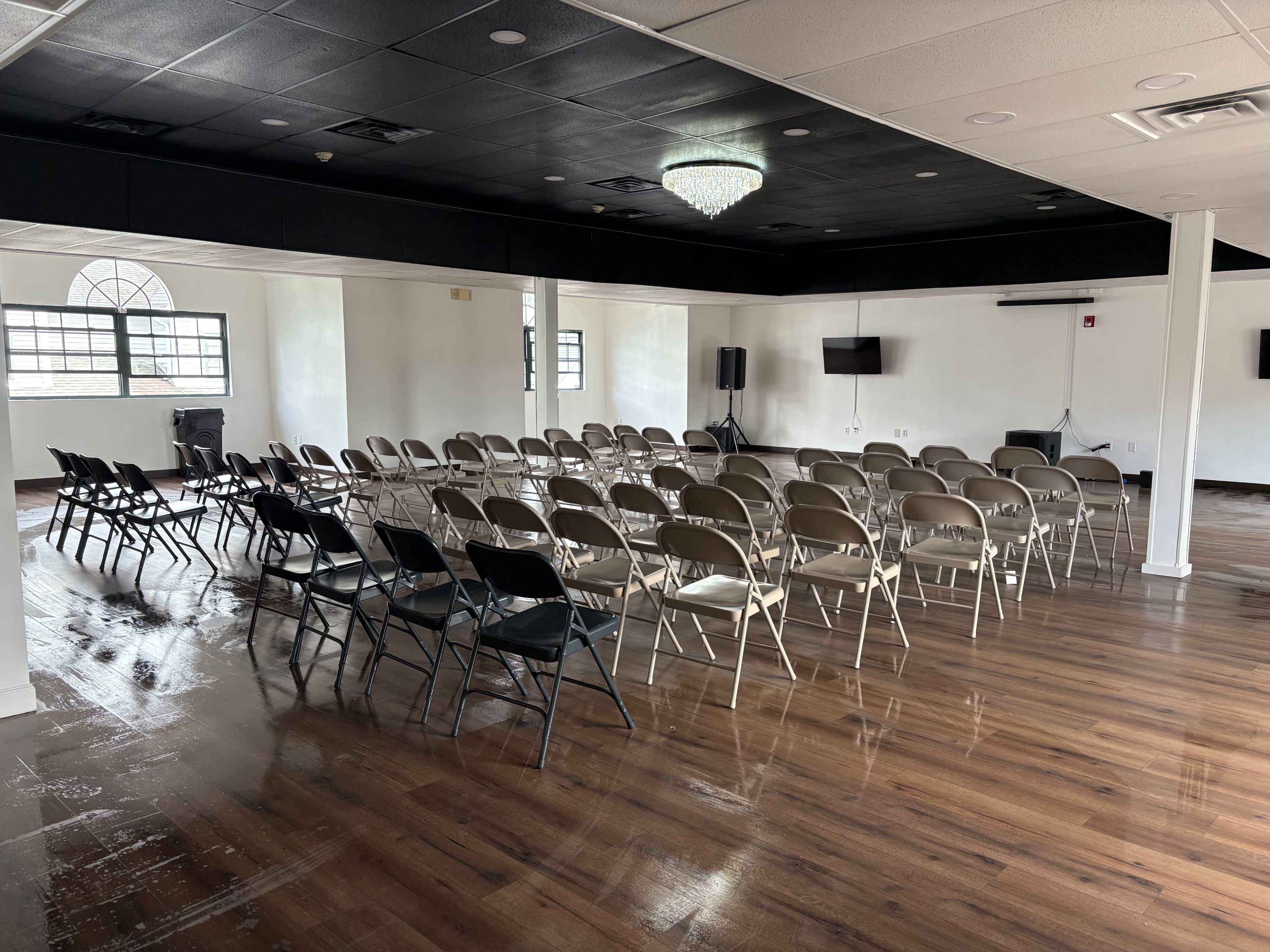 The image shows a spacious room with rows of folding chairs arranged facing a blank wall, illuminated by a chandelier overhead.