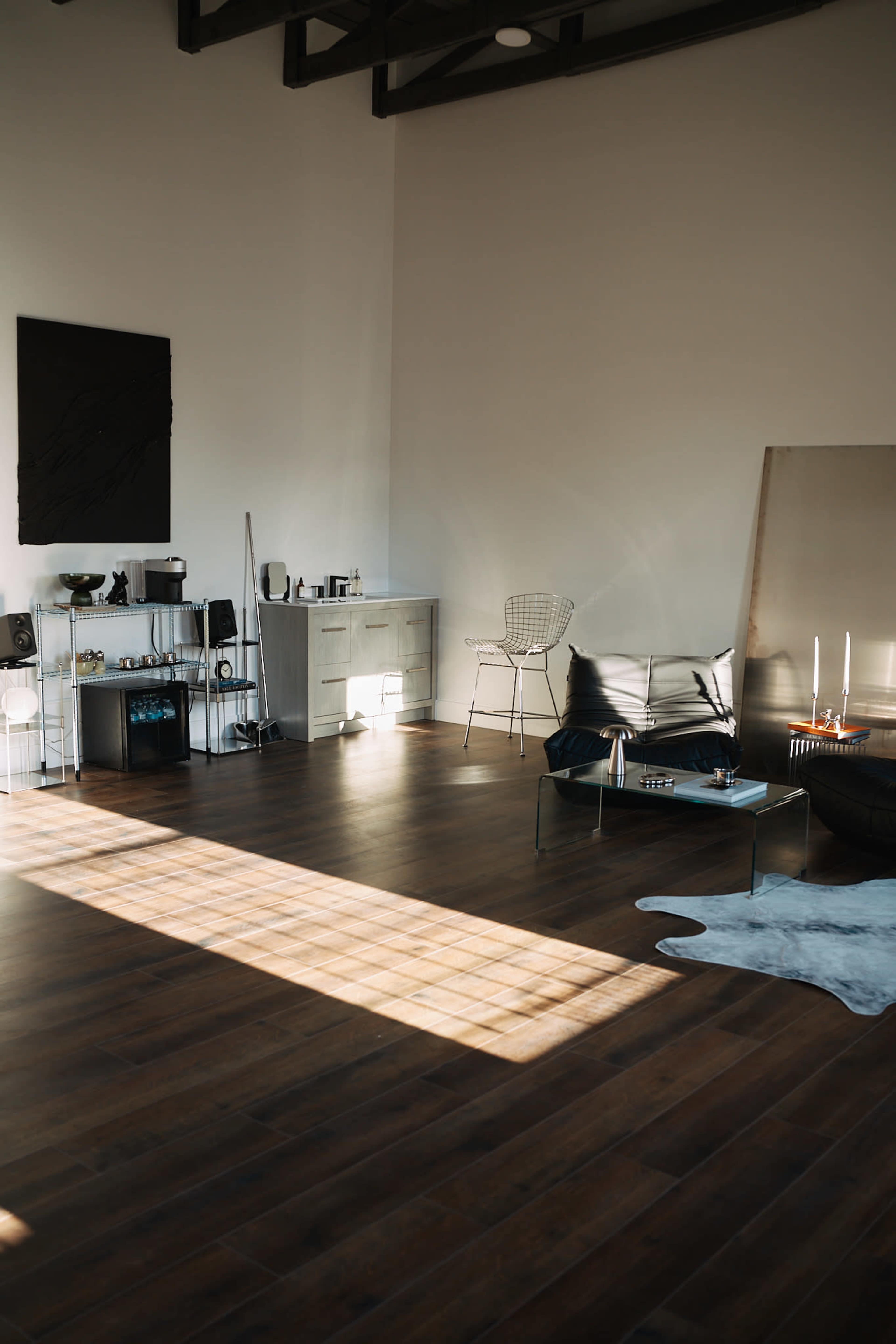 The image shows a spacious, well-lit living room featuring wooden flooring, a minimalist coffee table, a wire chair, and a sideboard against the wall.