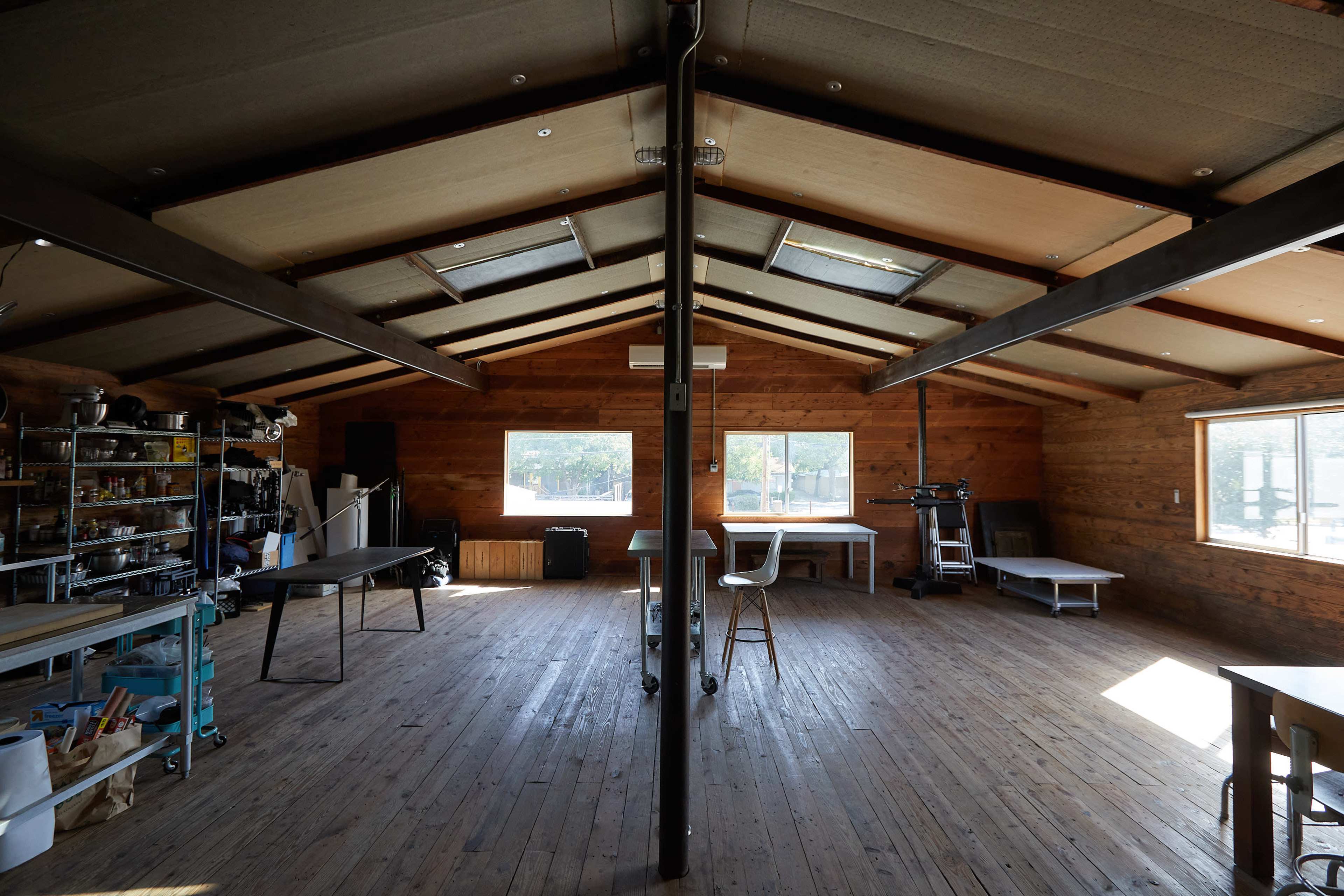 An open space with wooden walls, large windows, and various furniture arrangements including a table, chairs, and workstations.