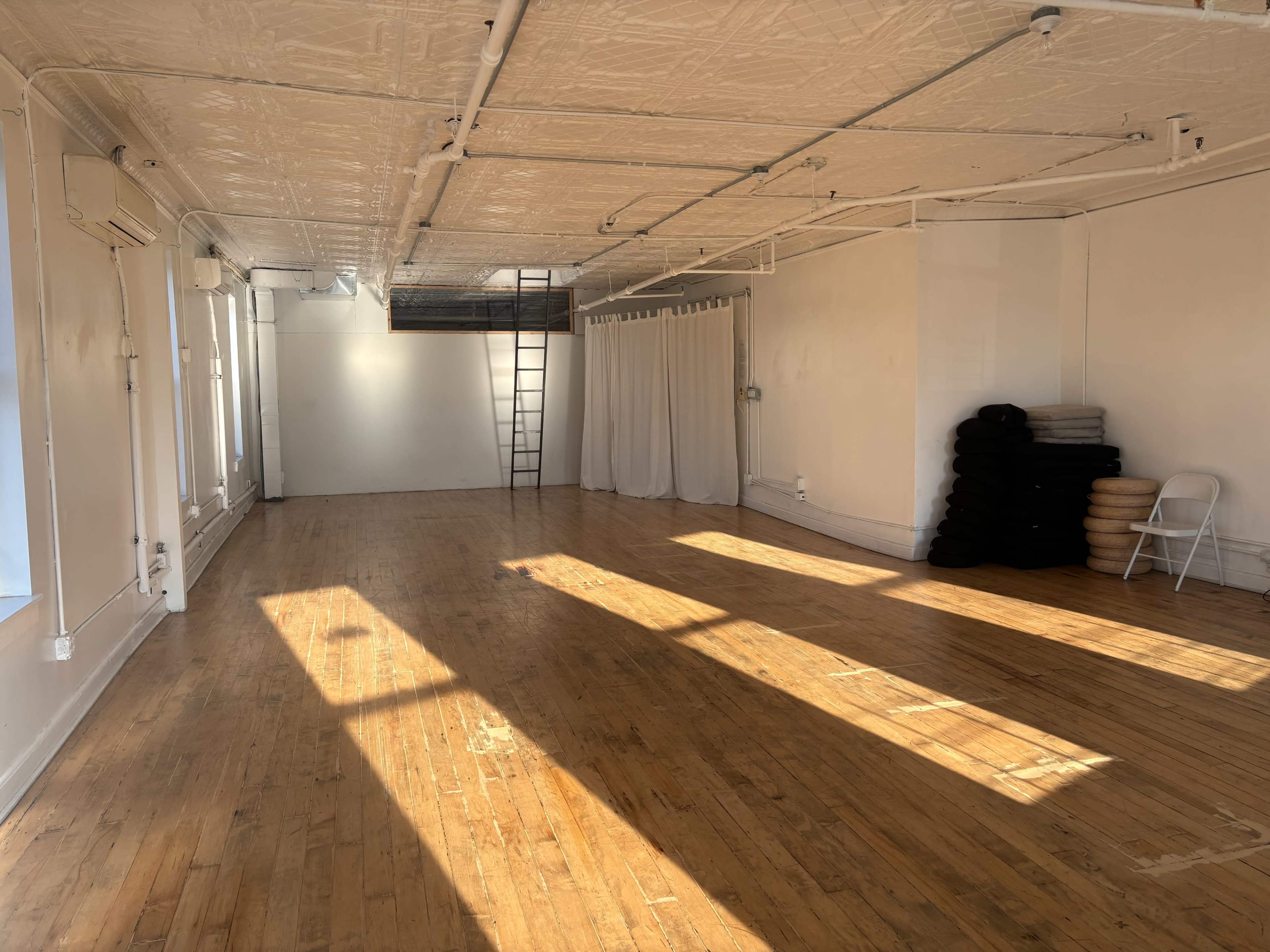 The image shows a spacious, empty wooden-floored studio with large windows allowing sunlight to stream in, a ladder, and neatly stacked cushions against one wall.